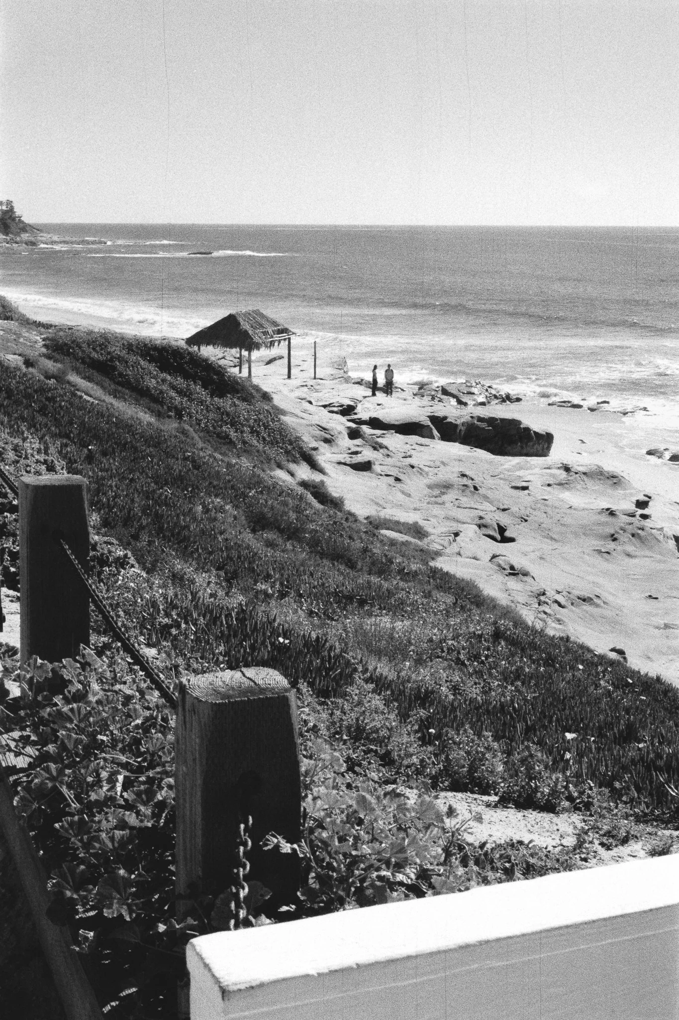Black-and-white coastal scene with rocky beach, ocean waves, thatched sun shelter, and two people by the shore