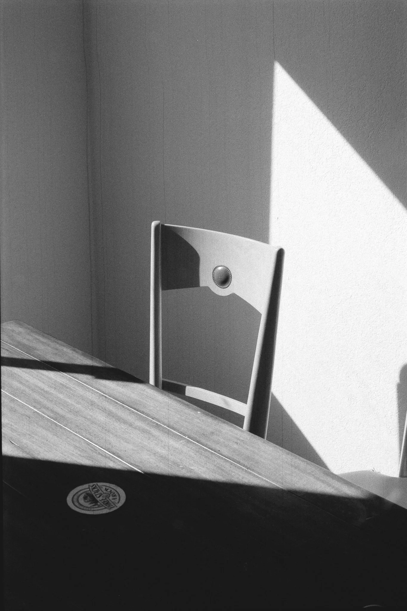 Black-and-white photo of an empty chair behind a wooden table, with strong sunlight casting geometric shadows on the wall.