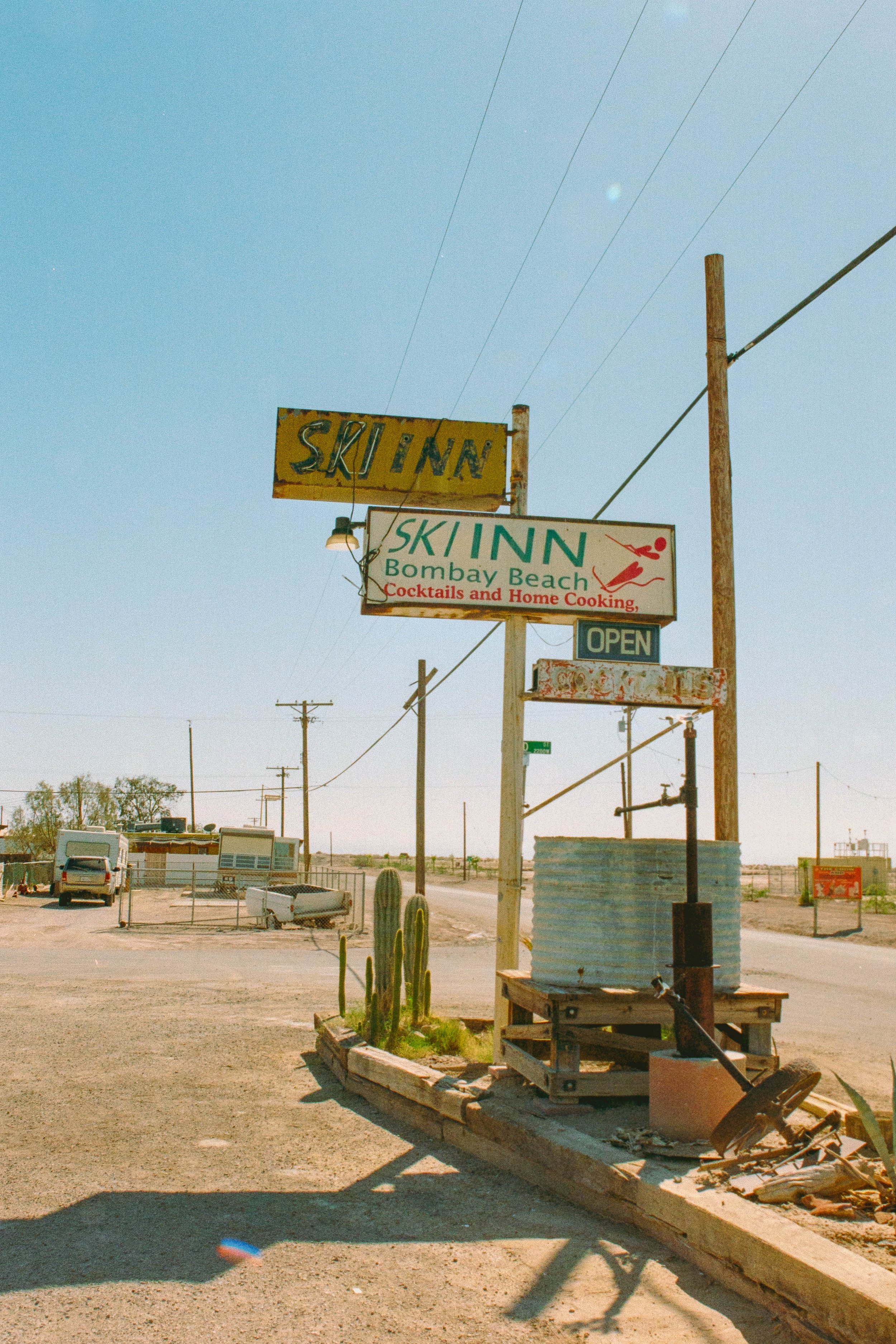 Weathered roadside signs for Sky Inn Bombay Beach in a desert landscape with utility poles, cacti, and an open sign.