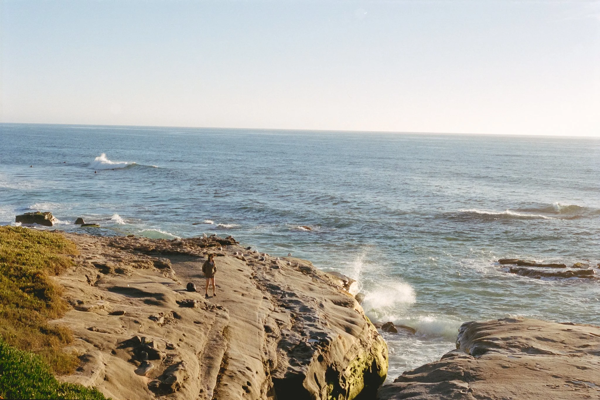 Rocky coastal cliffs overlooking ocean waves with surfers and a person standing on the shoreline