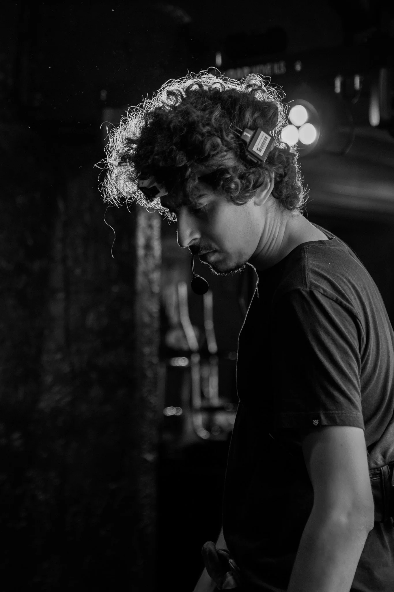 Black-and-white close-up of a curly-haired stage performer wearing a headset microphone and in-ear monitor.