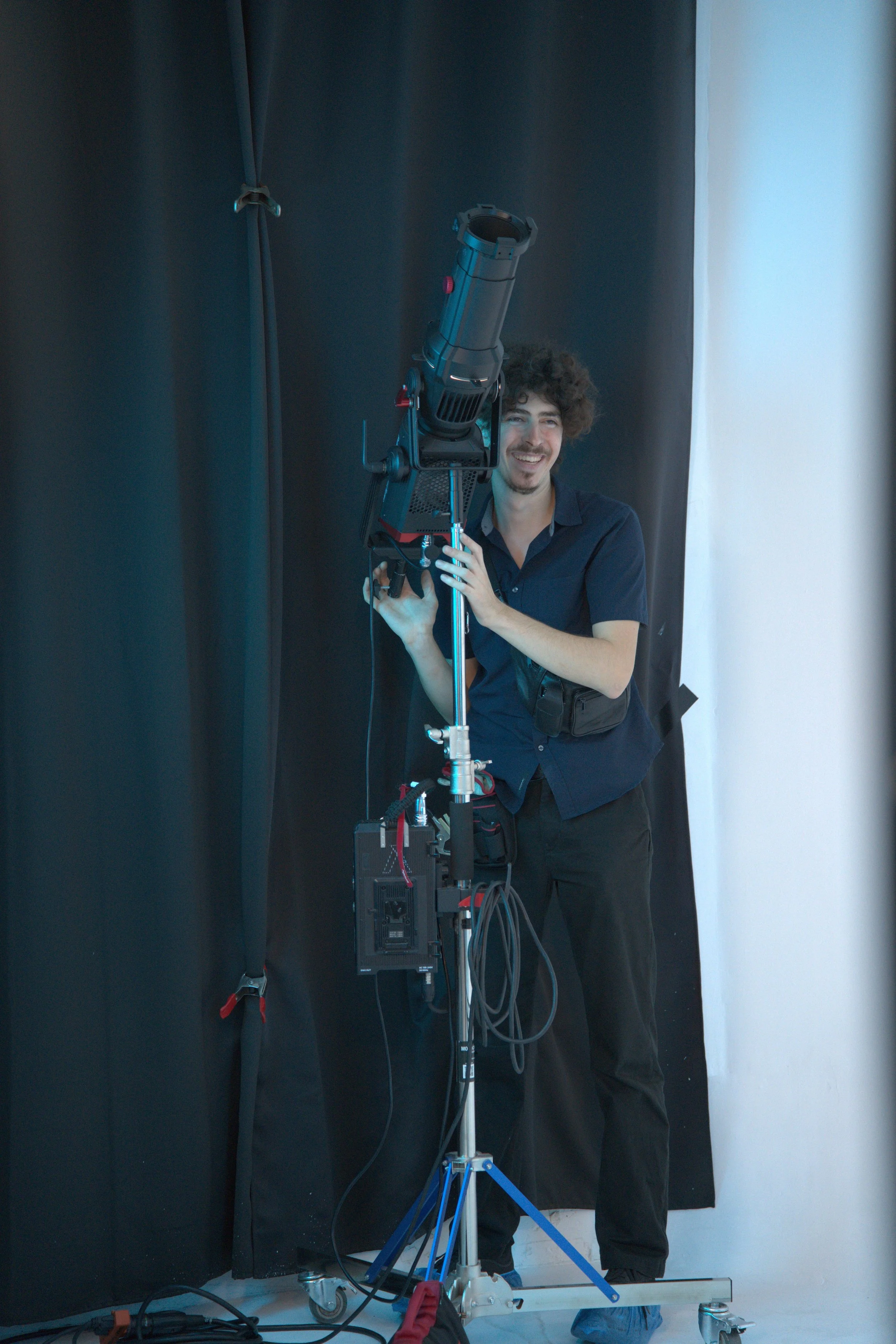 Smiling man operating a large studio spotlight on a stand in front of a black backdrop.