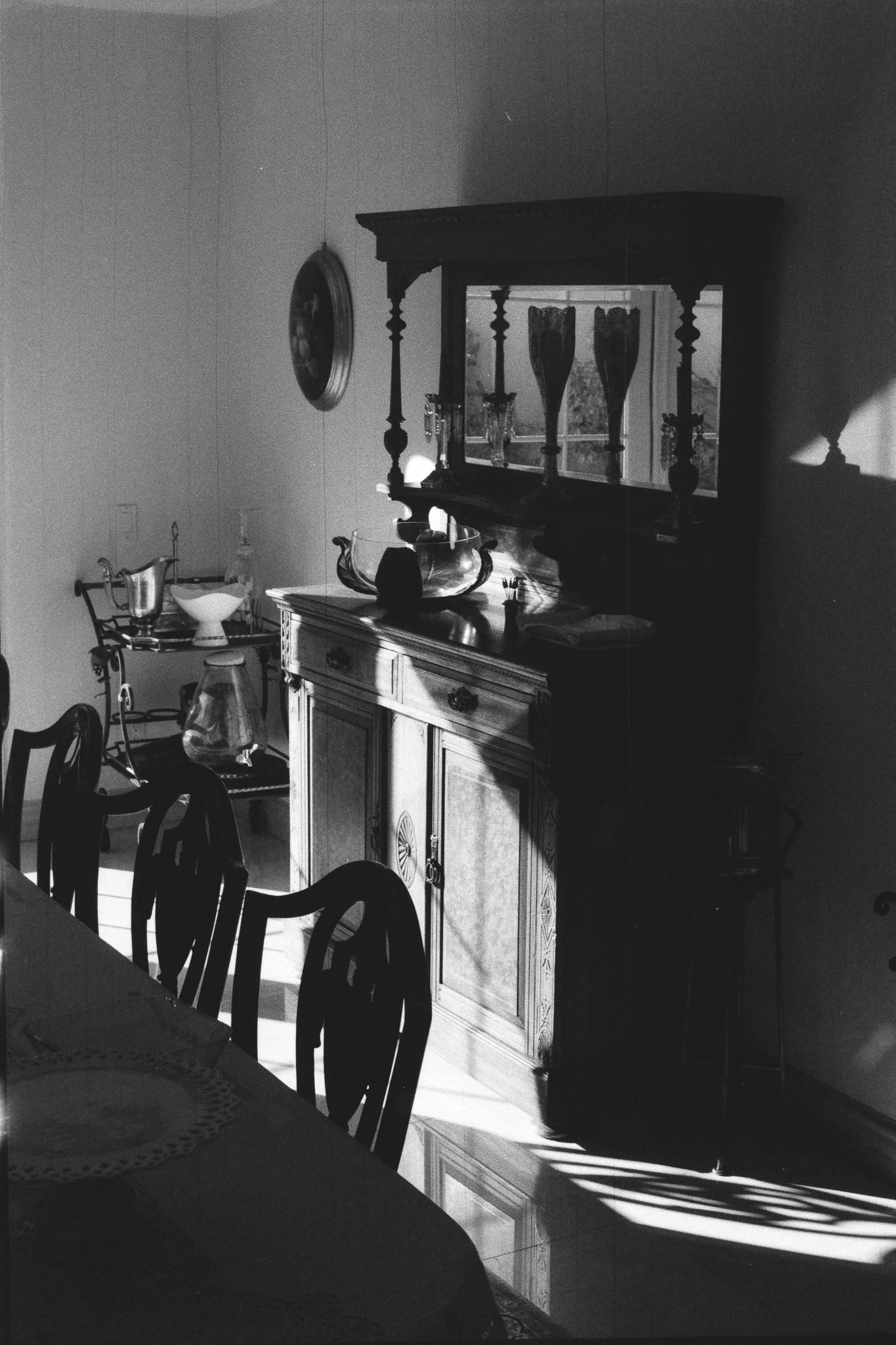 Black-and-white photo of a sunlit dining room with a carved wooden sideboard, large mirror, decorative vases, dining chairs, and table.