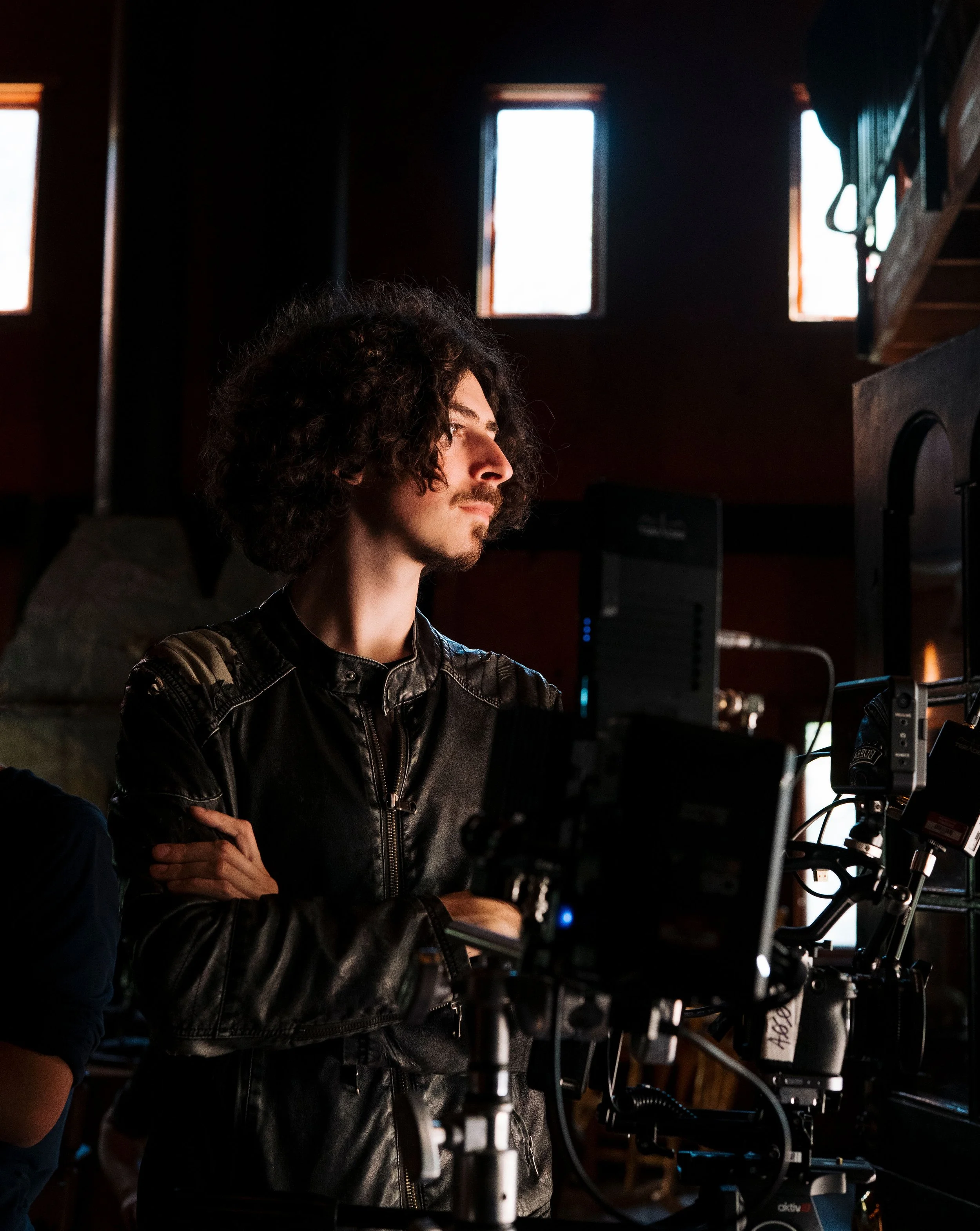 Curly-haired man in a black leather jacket standing on a film set beside professional camera equipment