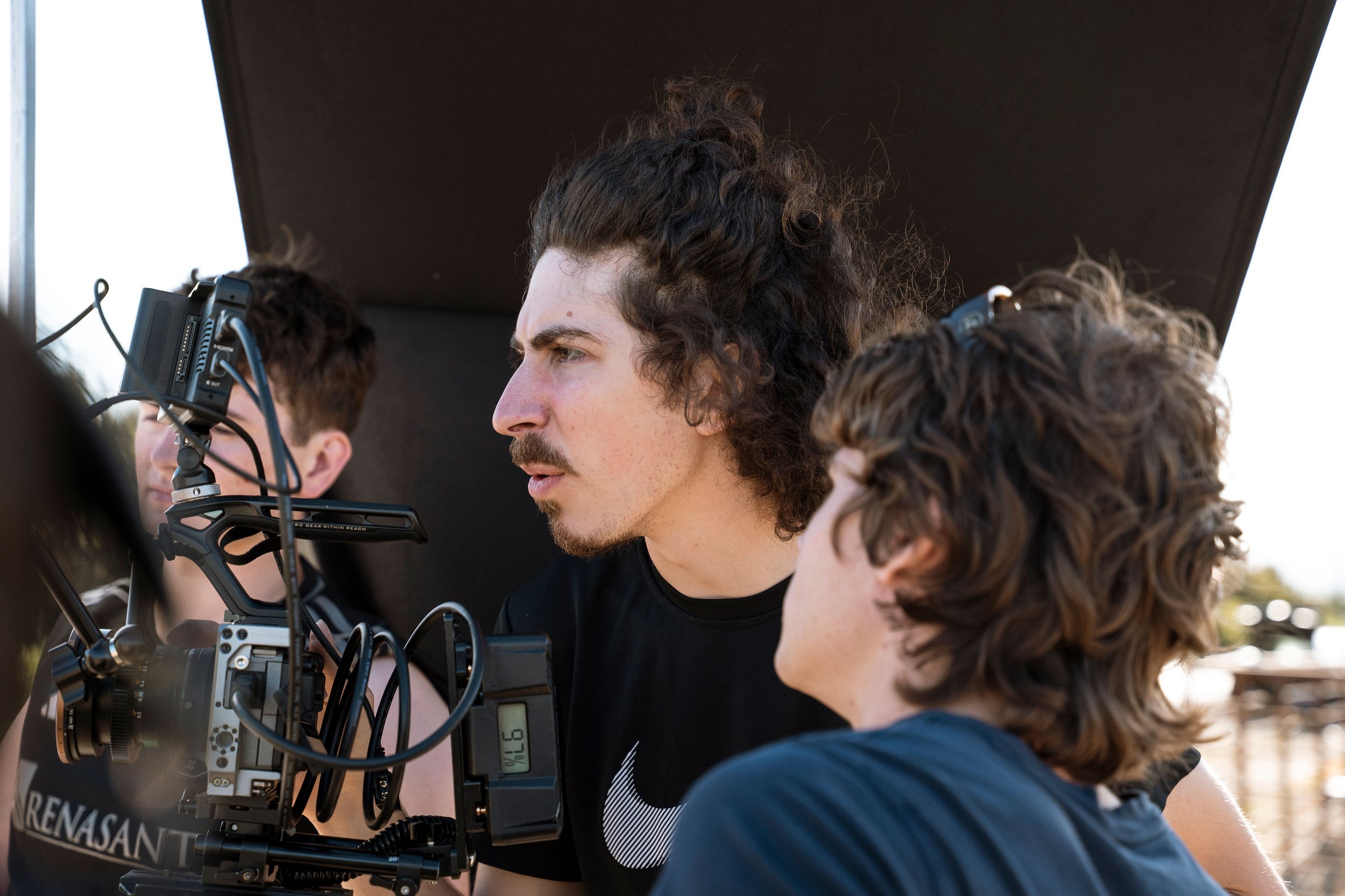 Film crew operating a professional cinema camera outdoors with a curly-haired man in the center.
