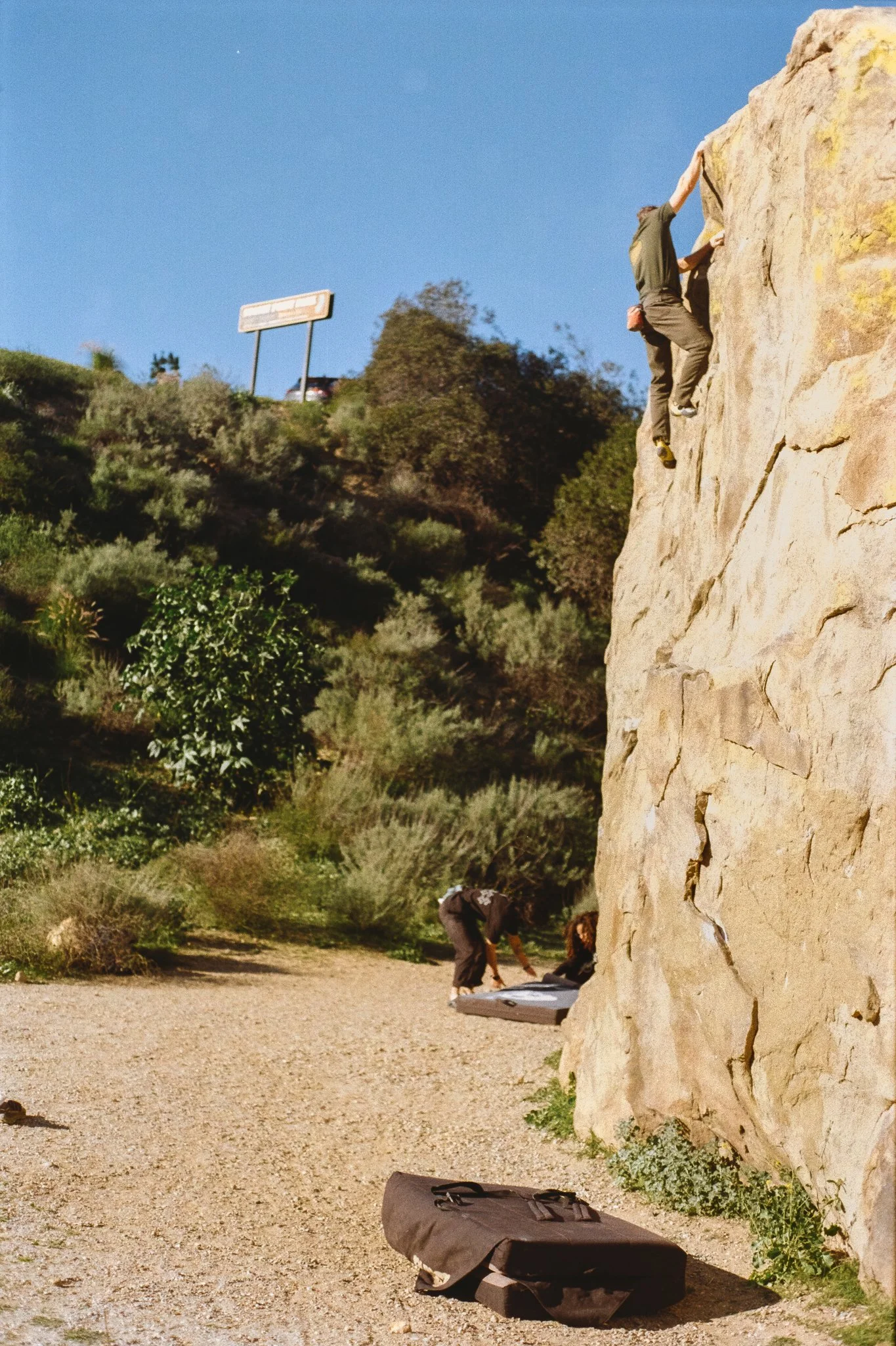 Rock climber scaling a sunlit sandstone cliff with crash pads below