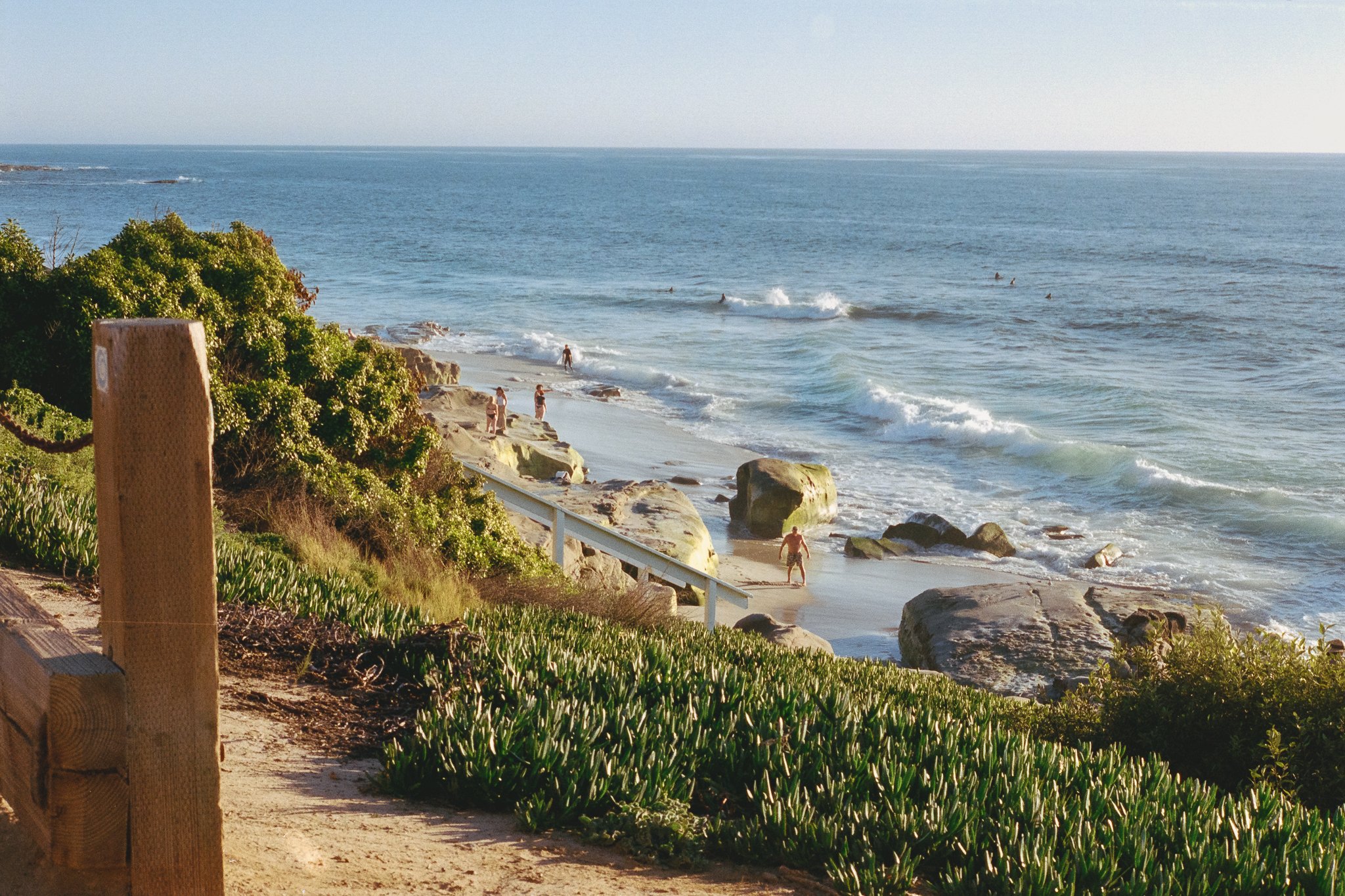 Rocky ocean beach with surfers, swimmers, coastal vegetation, and a wooden fence overlooking the waves