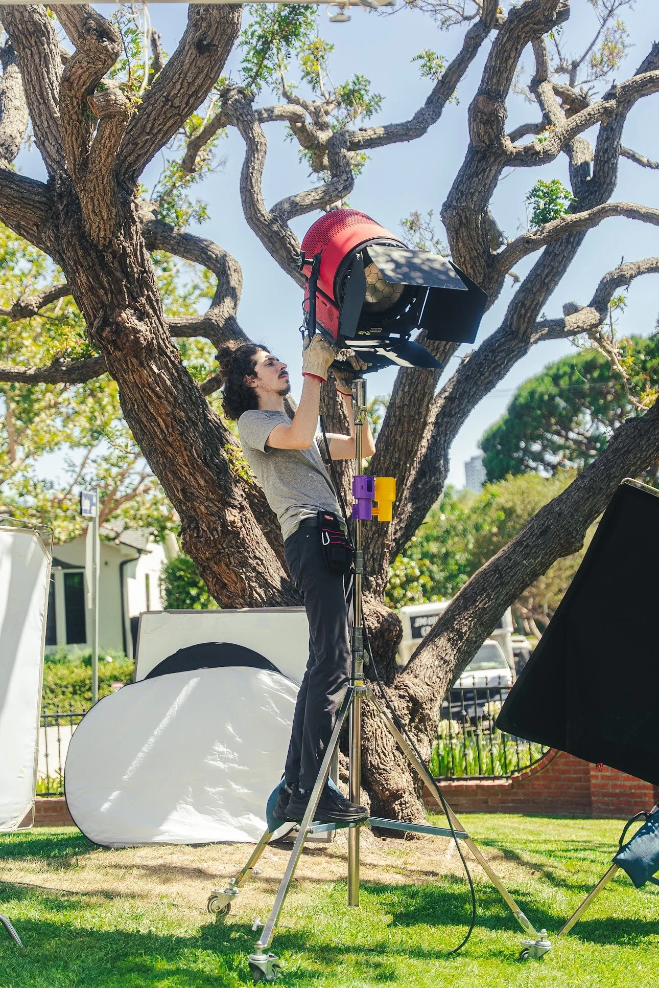 Film crew member operating a large studio light on a rolling stand outdoors beneath a tree
