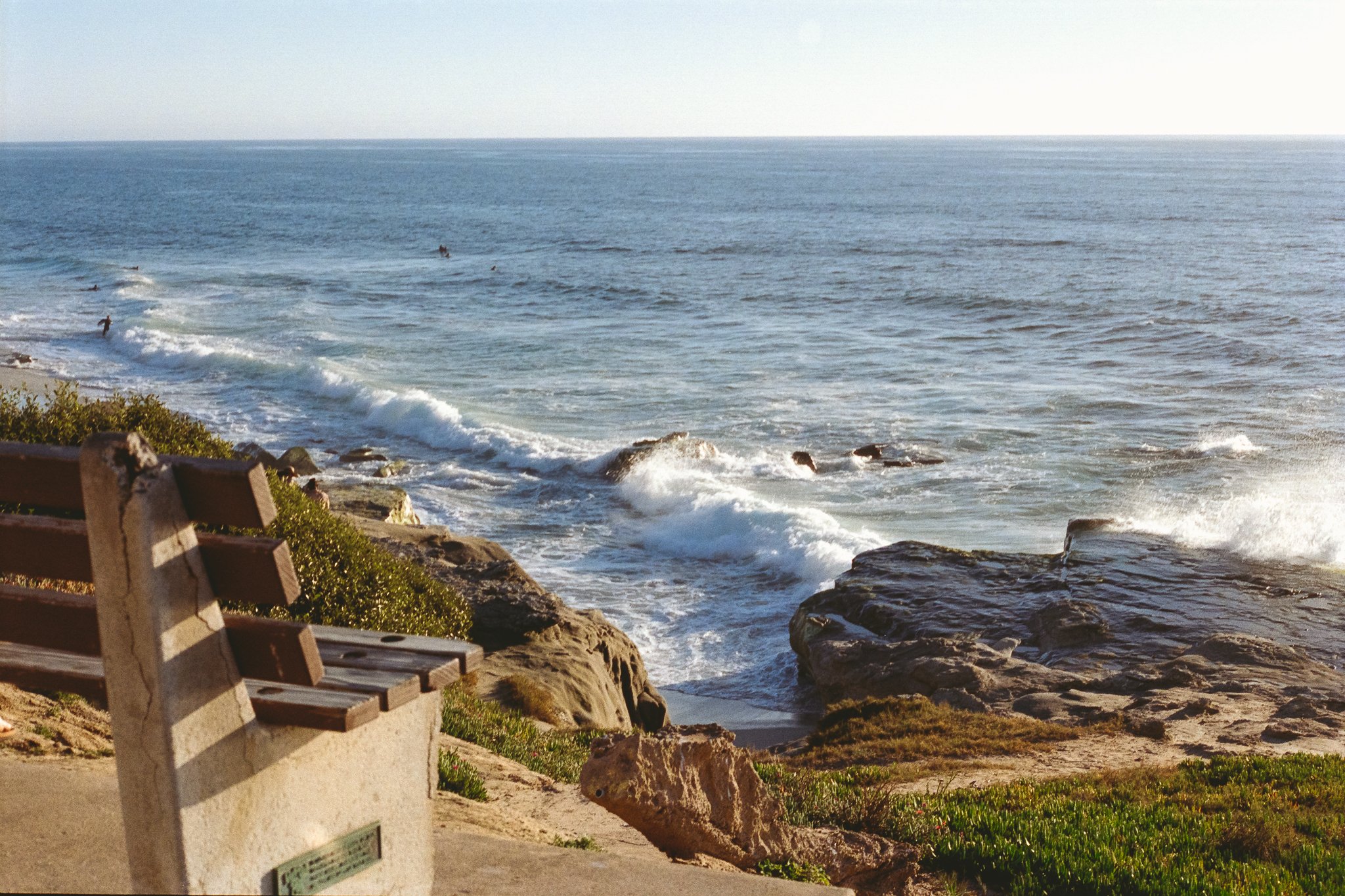 Rocky ocean coastline with waves, surfers, and a wooden bench overlooking the water