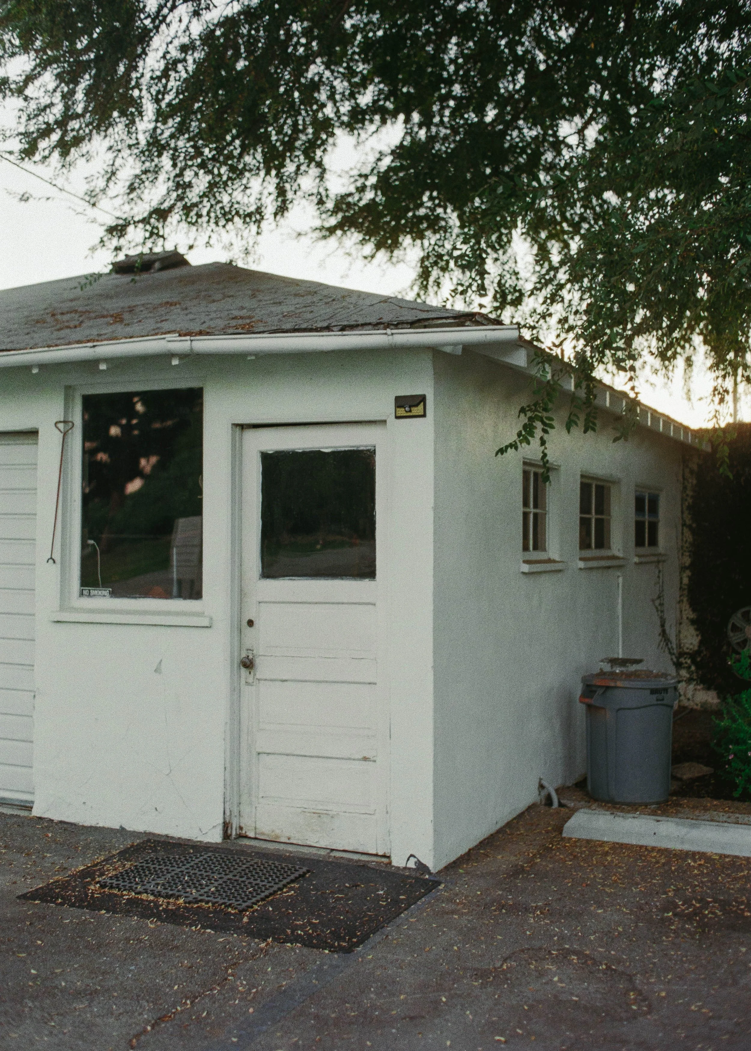 White garage with windows and doors beneath leafy trees, featuring a trash bin and garden tools outside.