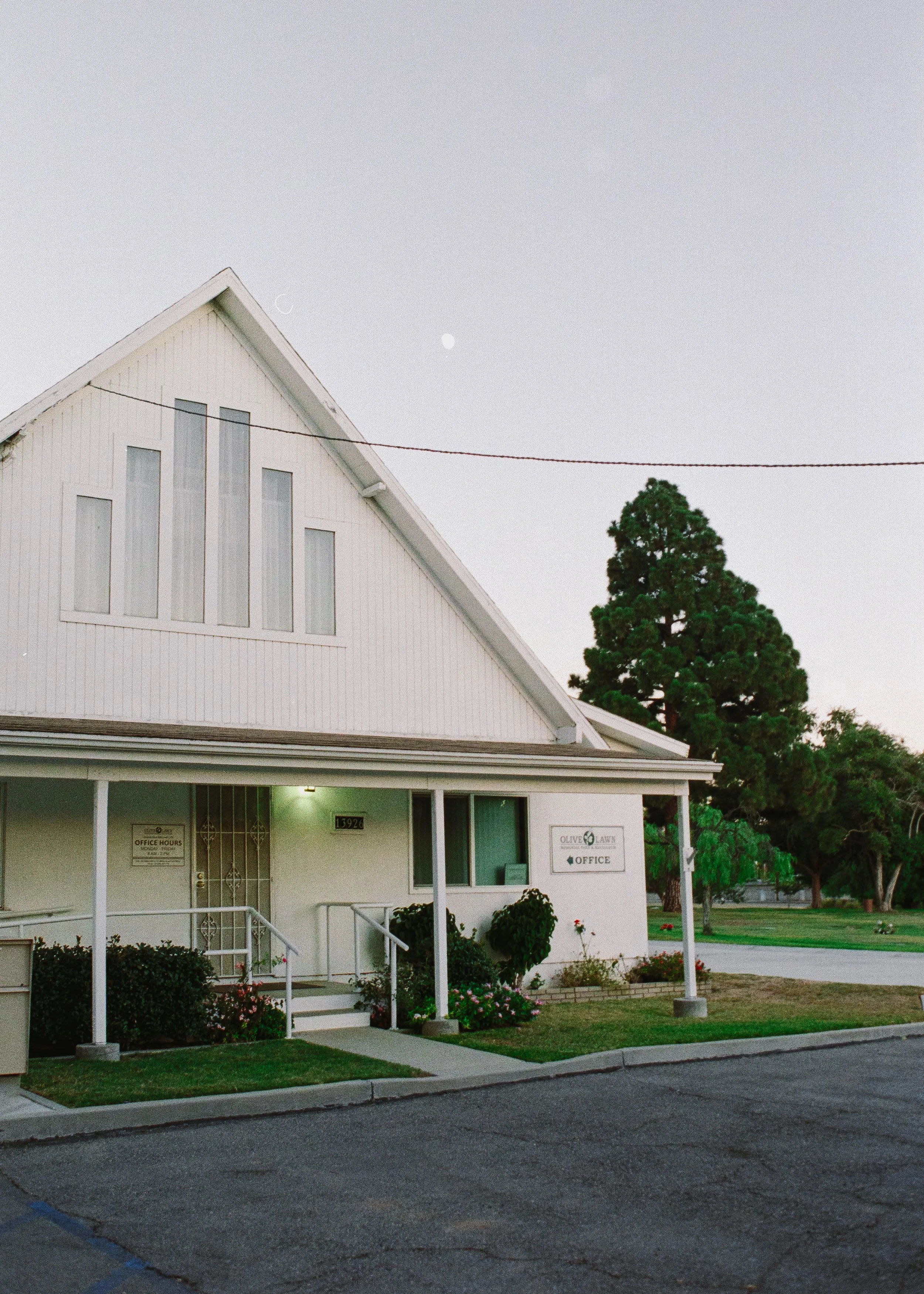 White church office building with a peaked roof, porch, flower beds, and large evergreen tree