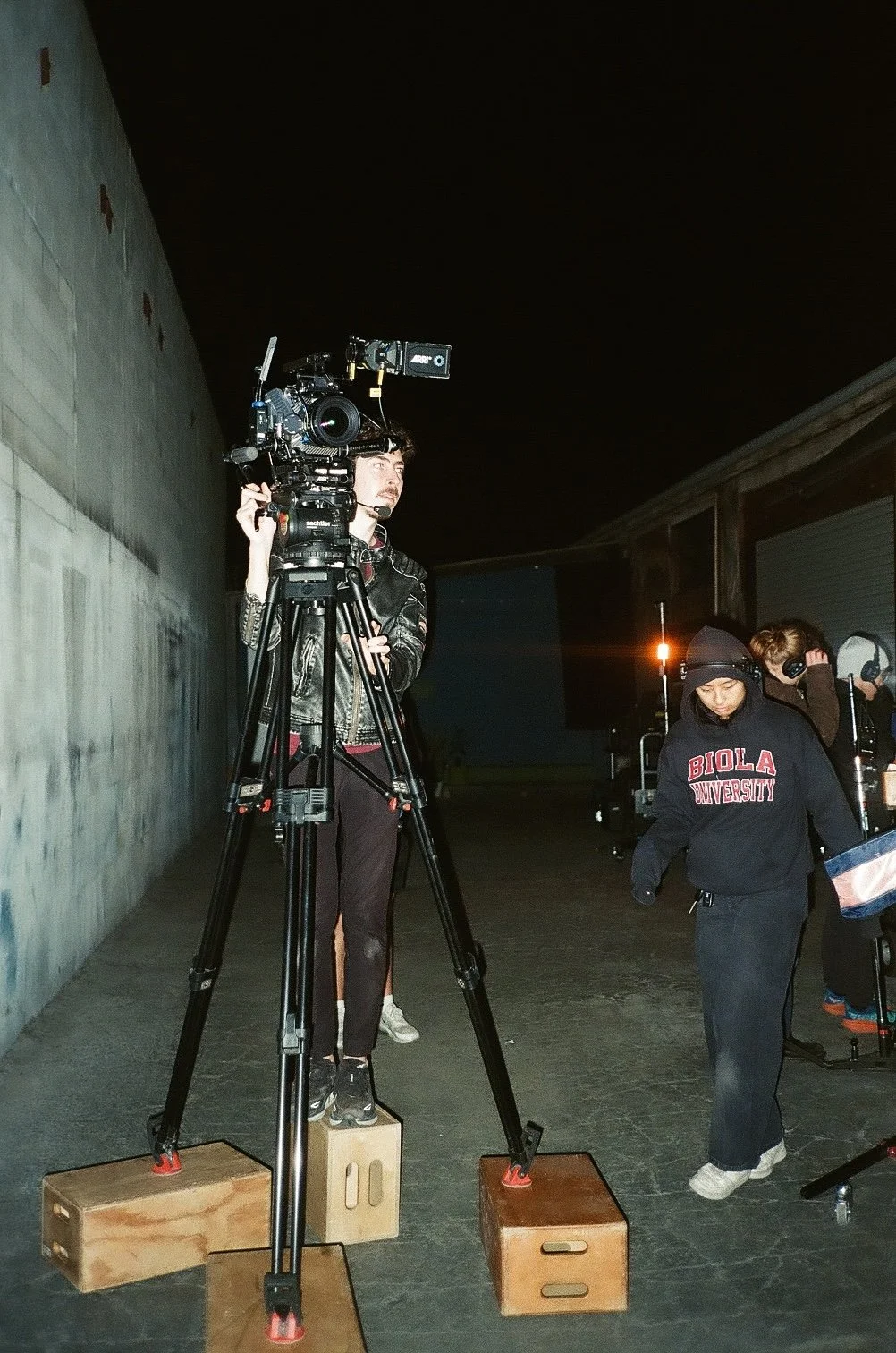 Film crew filming at night with a professional camera on a tripod and crew members in a studio alley.