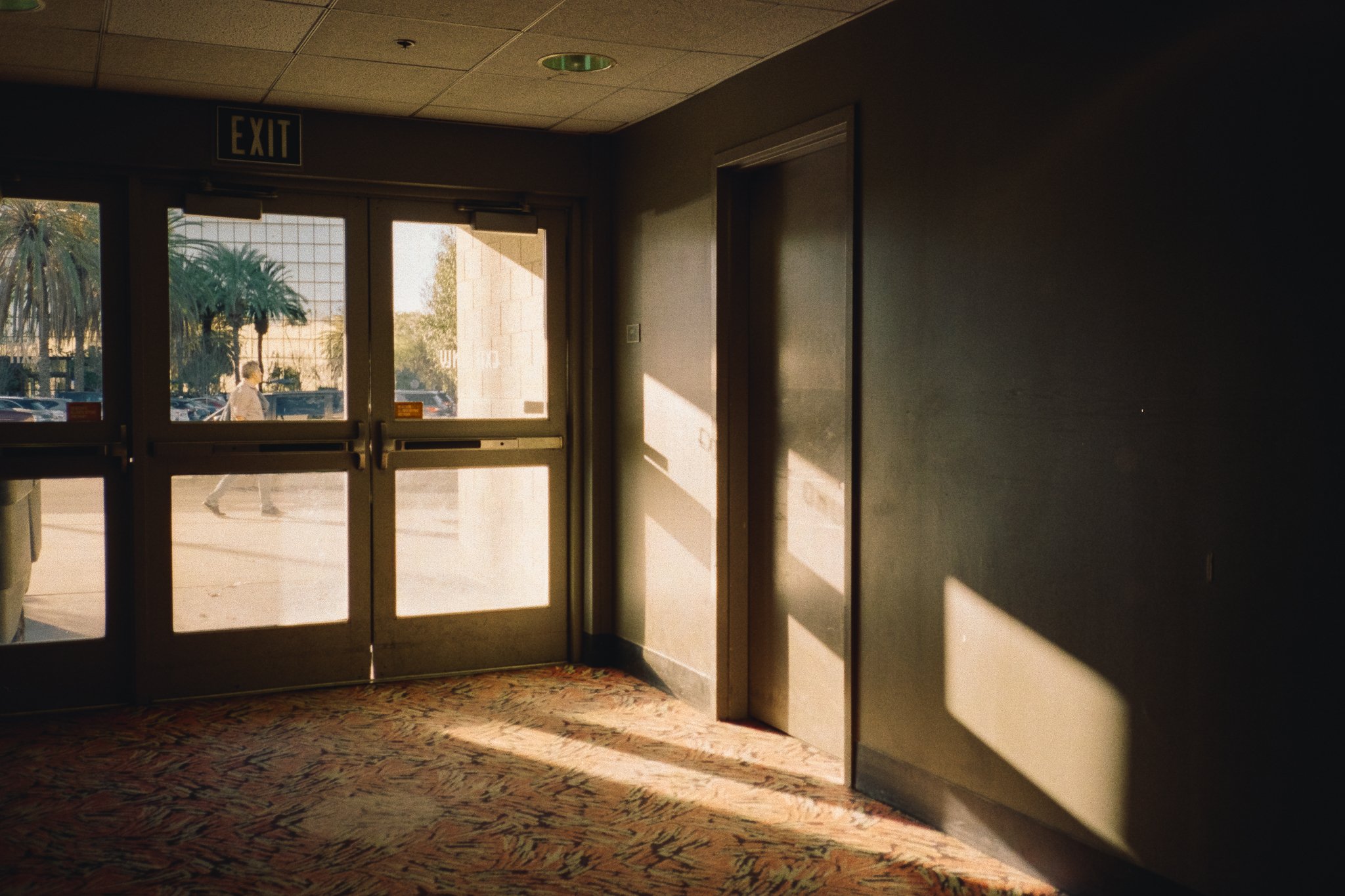 Sunlit building lobby with glass exit doors, patterned carpet, and a person walking outside