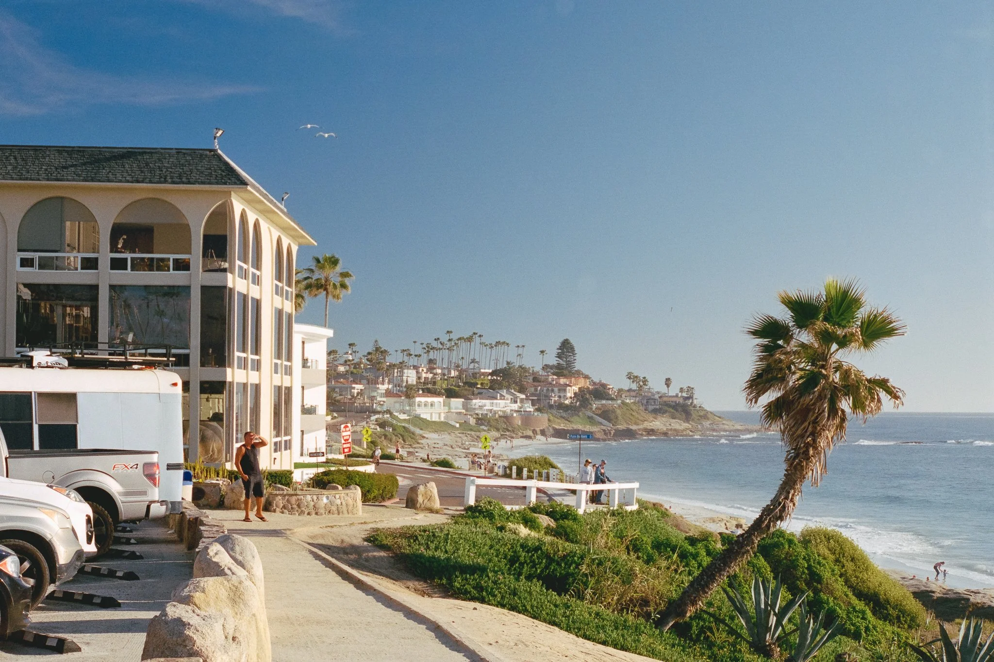 Coastal California beach neighborhood with oceanfront buildings, palm trees, parked vehicles, and a sunny blue sky