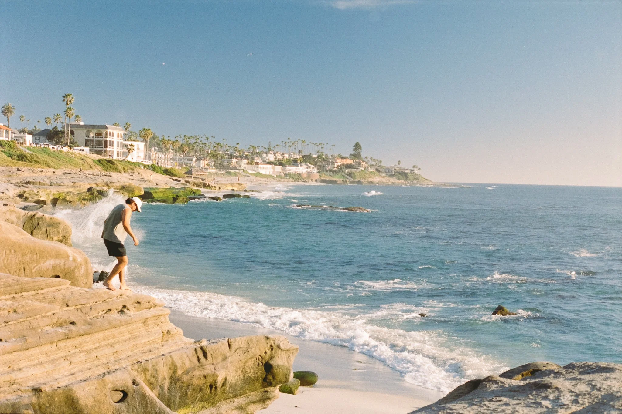 Person standing on rocky coastal cliffs overlooking ocean waves, sandy beach, and palm-lined seaside homes