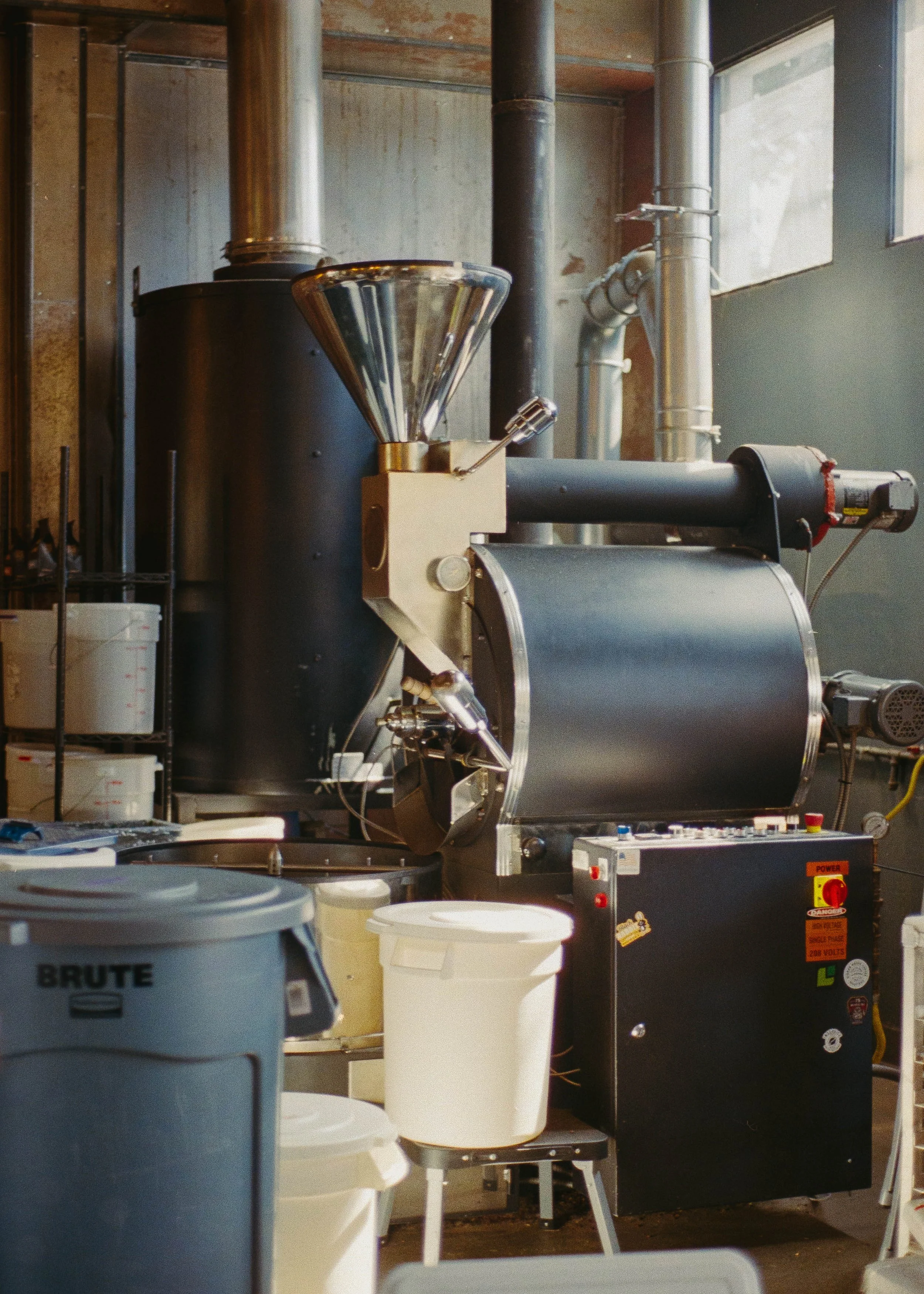 Industrial workshop interior with large pipes, tanks, hoses, plastic buckets, and processing equipment.