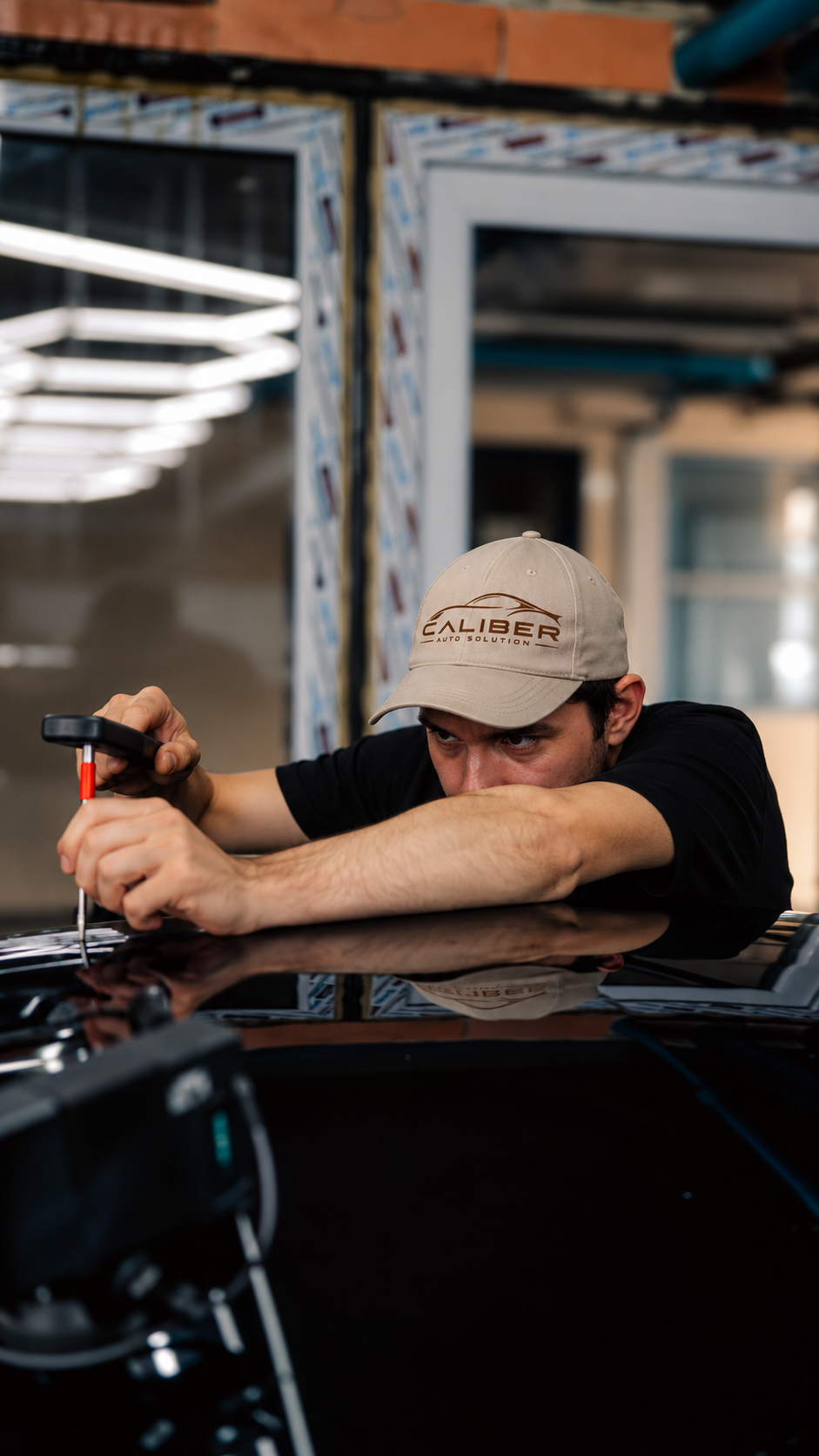 A man wearing a beige cap with the logo 'Caliber Auto Solution' is fixing a car using a screwdriver in an indoor workshop. He is leaning on the car hood, focused on his work.