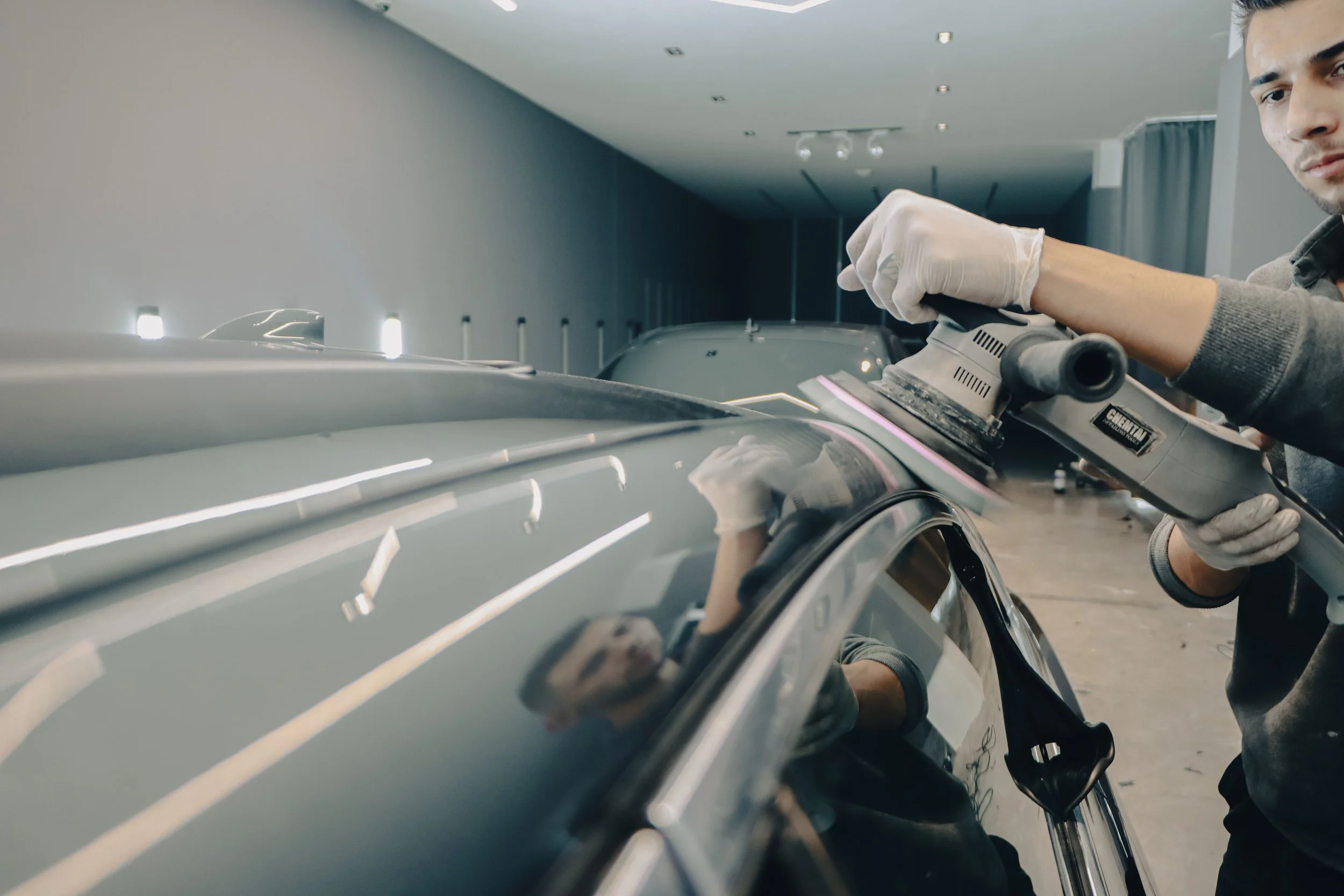 A man in gloves is using a power sander on a car in a workshop. The car is light-colored and shiny, and the man is focused on his work.