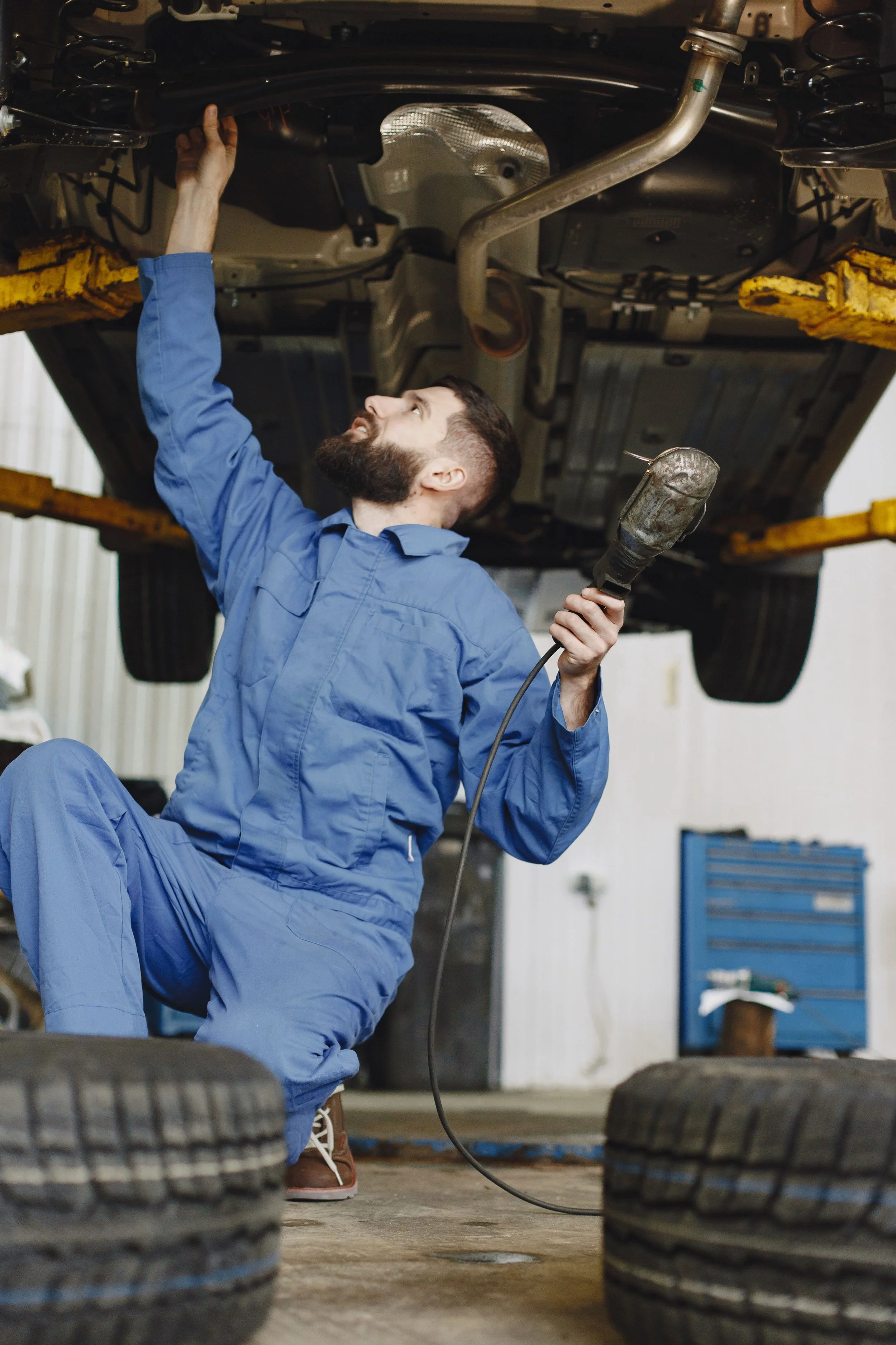 A mechanic working underneath a car, using a flashlight and inspecting or repairing the vehicle, with tires in the foreground and tools and equipment in the background.