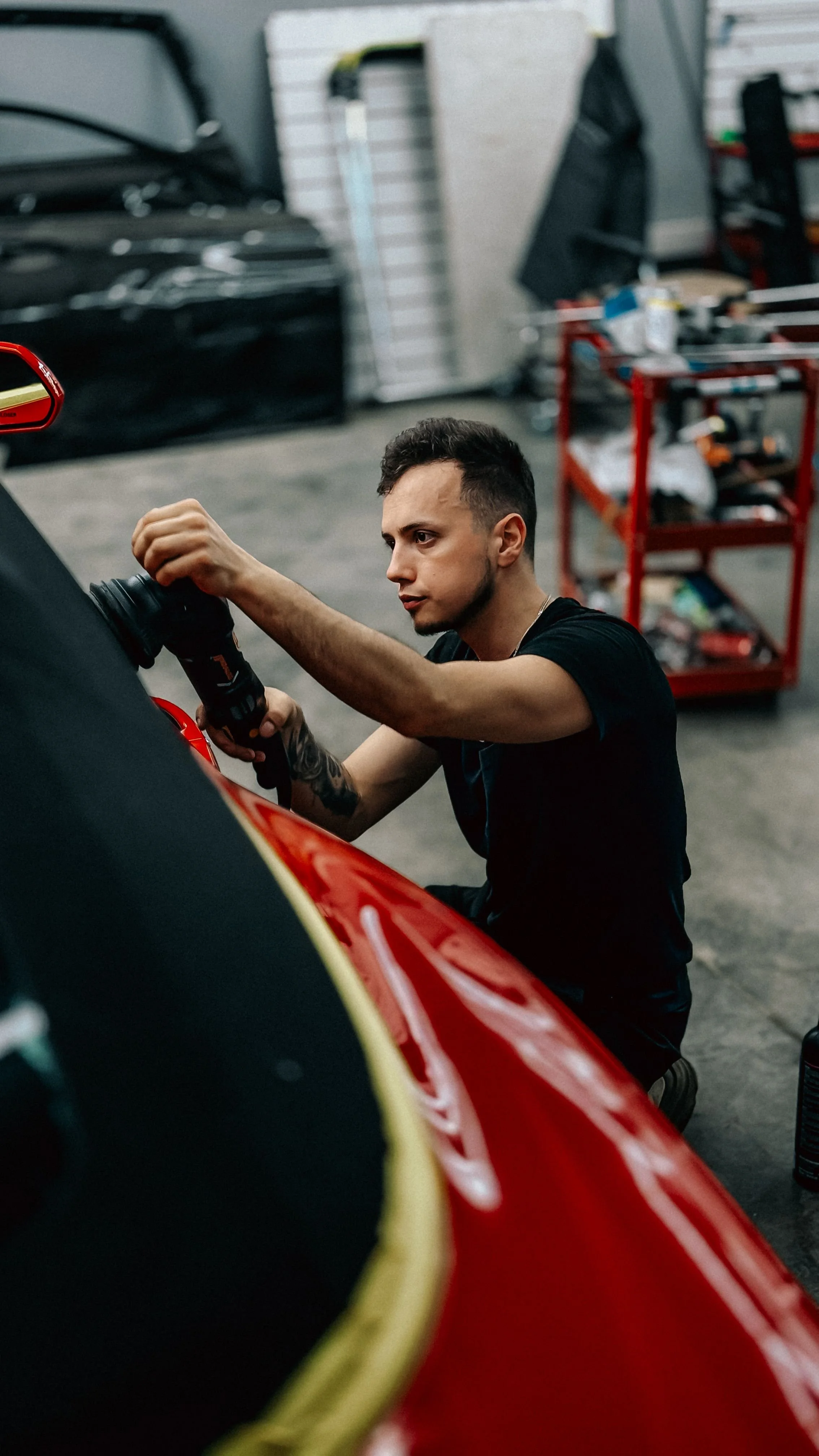 Young man working on a red sports car in a garage, using a power tool.