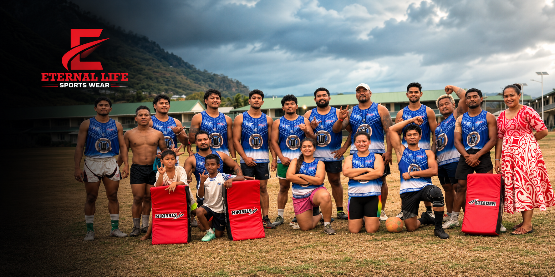 A group of men and women wearing blue sports jerseys with a logo, posing on a field with a mountainous background. Two children sit on the ground in front, and a woman in a red and white dress stands on the far right. Some team members are making victory signs or flexing muscles. The logo 'Eternal Life Sports Wear' appears in the top left corner.