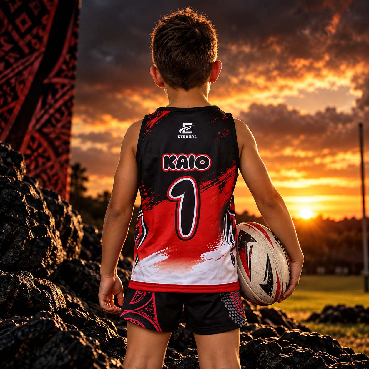 A young boy stands outdoors at sunset, wearing a black and red sports jersey with the number 1 and the name KAIO on the back, holding a rugby ball, with dark rocks and a field in the background.