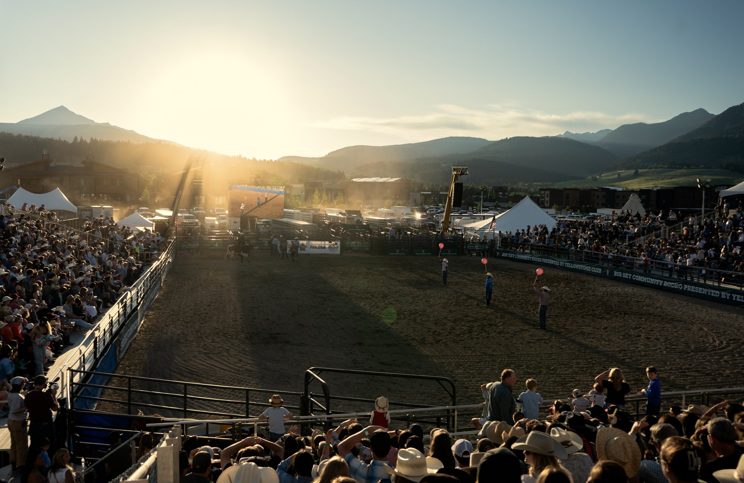 Crowd seated in stands watching a rodeo arena at sunset with mountain scenery in the background.