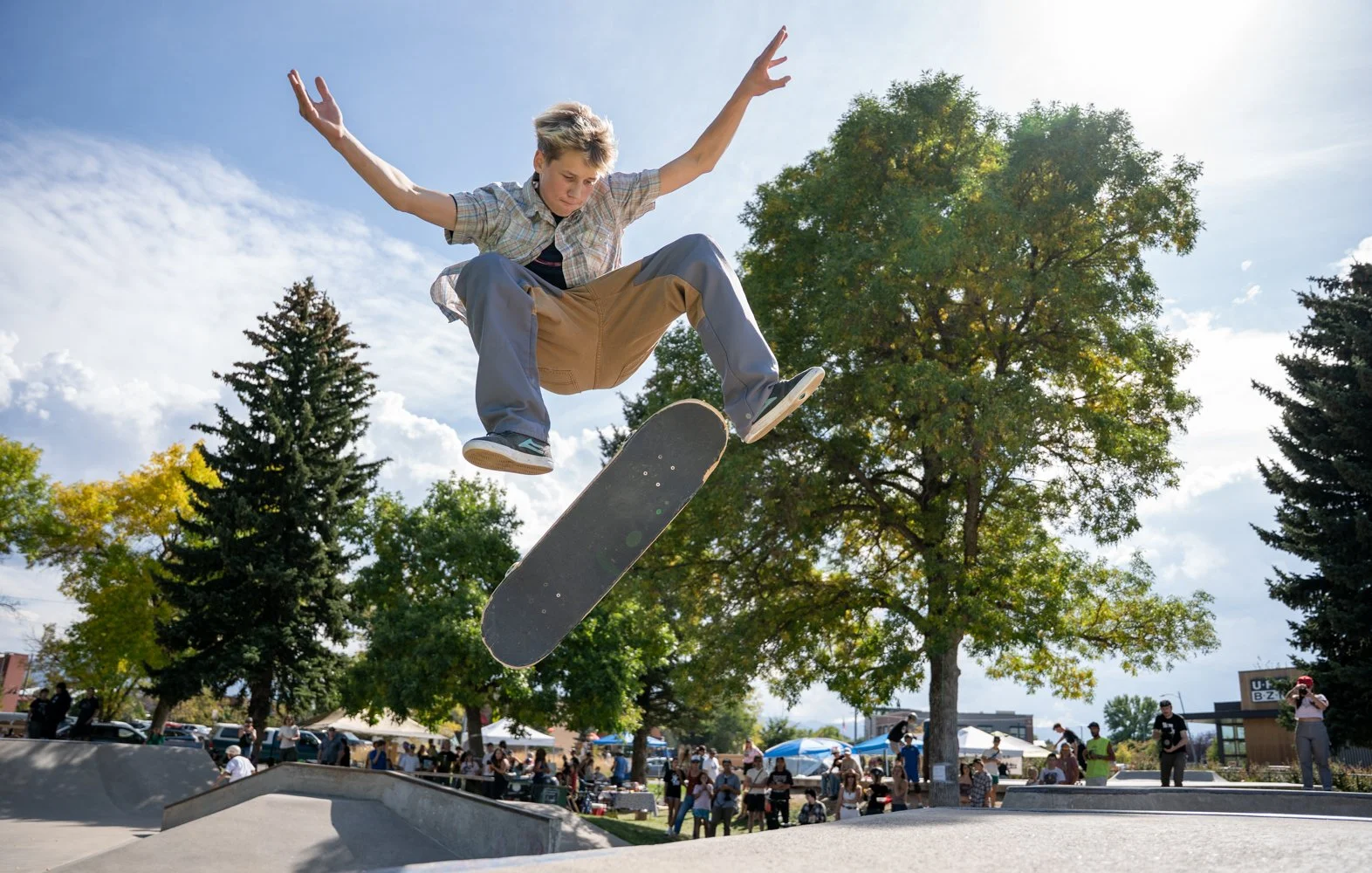 Skateboarder performing an ollie at a skate park on a sunny day, with spectators and trees in the background.