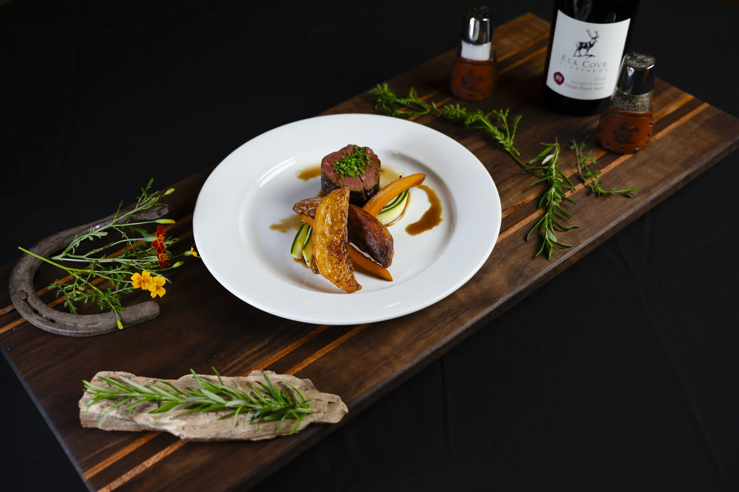 A white plate with cooked beef tenderloin topped with green herbs, served with roasted vegetables and sauce, on a wooden serving board decorated with sprigs of rosemary and greenery, alongside bottles of wine and spice containers.