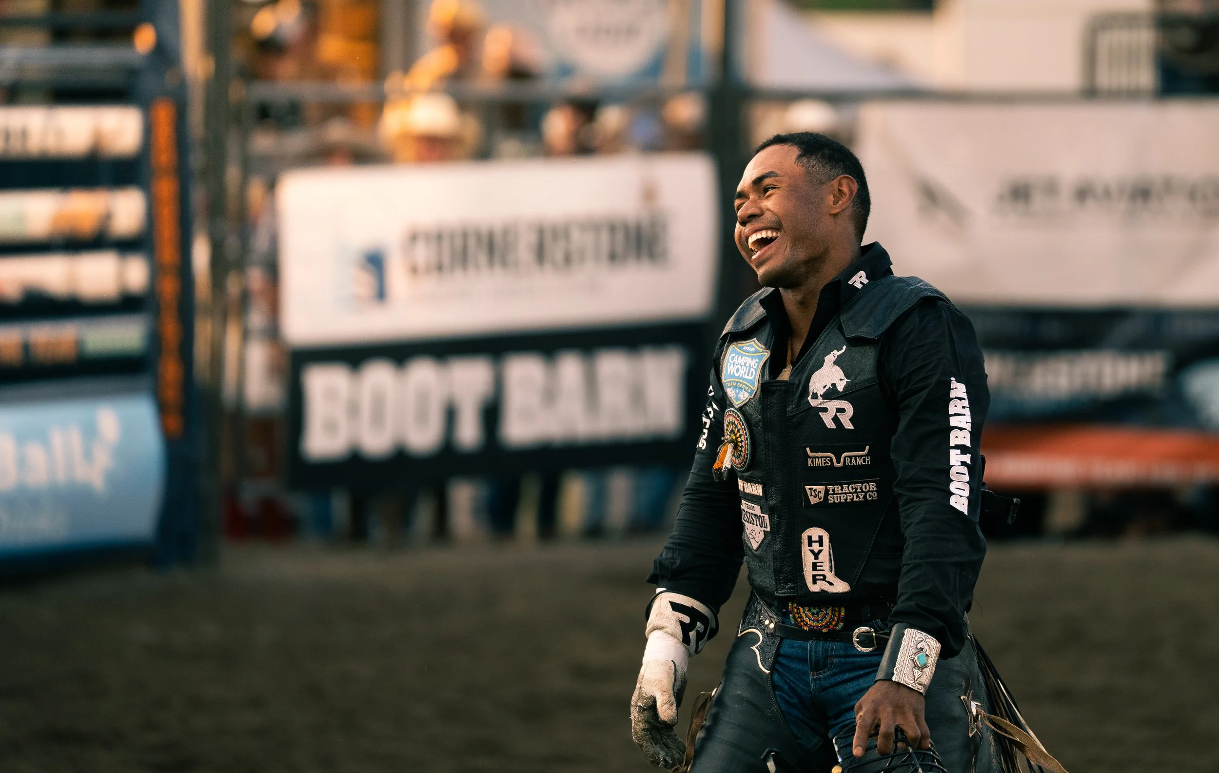 A man in a black racing suit with various patches and logos is smiling and holding a helmet in his left hand, standing outdoors with a blurred background of signs and structures.