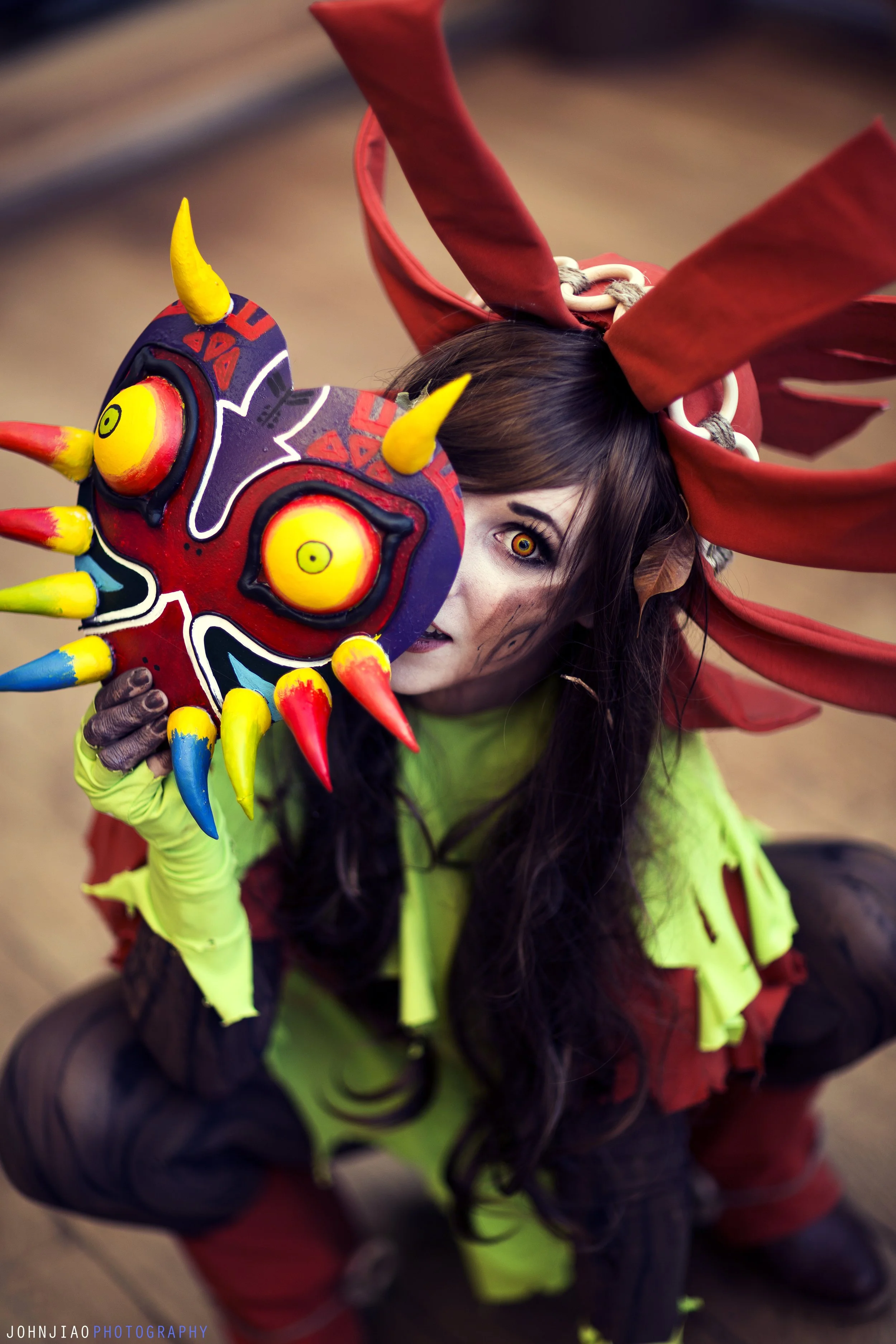 A person dressed in a vibrant, elaborate costume inspired by traditional indigenous or mythological attire. They hold a colorful, stylized mask with striking yellow eyes, red, yellow, blue, and purple accents, and spiked protrusions, partially covering their face. The costume includes red and brown headgear with large, pointed, leaf-like extensions, a bright neon green garment with black accents, and additional costume details. The scene appears to be indoors, possibly at a cultural or artistic event.