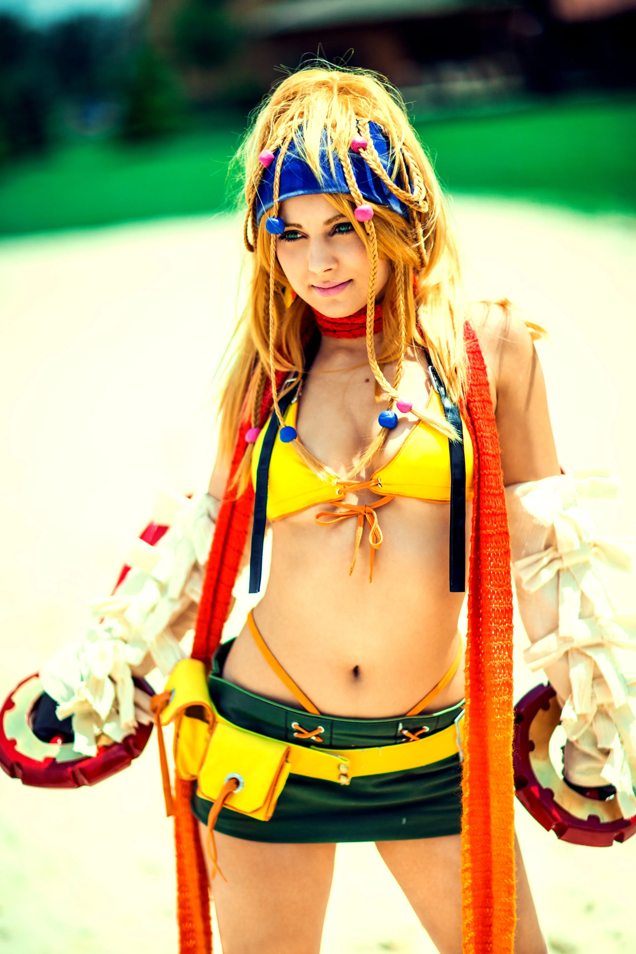 A young woman with blonde dreadlocks wearing a blue bandana, yellow bikini top, black shorts with orange accents, and holding climbing gear, standing outdoors with a green background.