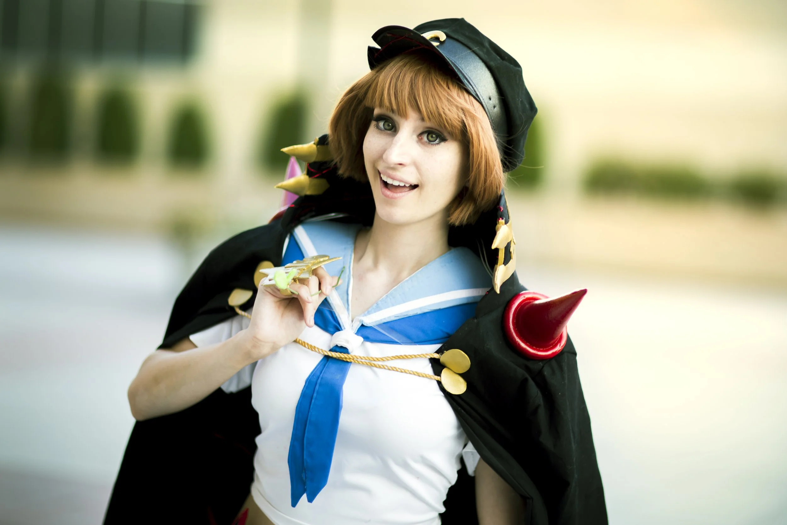 Young woman dressed in cosplay costume with short reddish hair, eye makeup, sailor-style top, and a black cape with colorful spikes accessory.