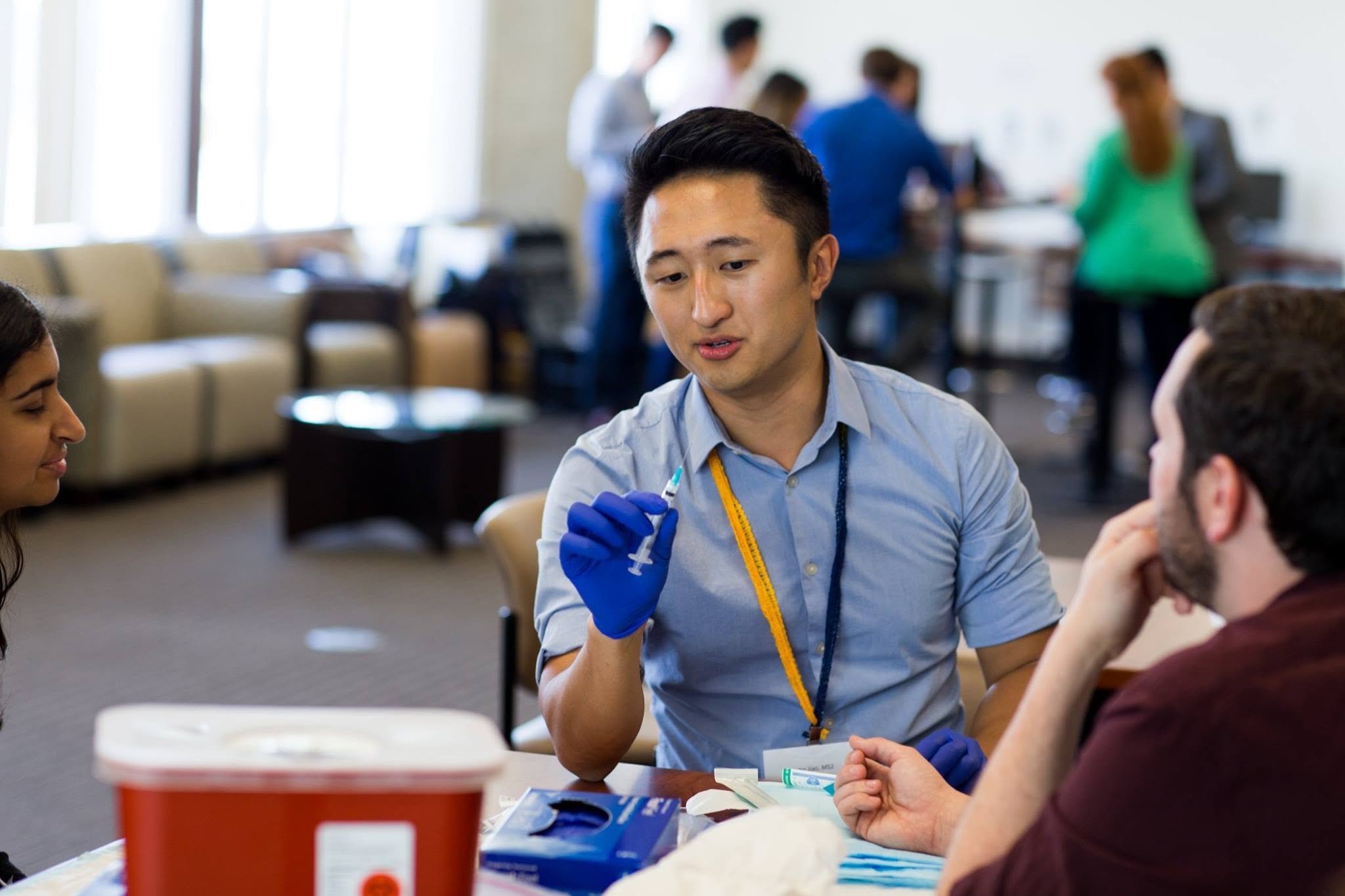 A young man with black hair, wearing a light blue shirt and blue gloves, is preparing a syringe in a clinical setting. He is seated at a table with medical supplies, and several people are in the background, some working at tables and others standing, in a well-lit room.