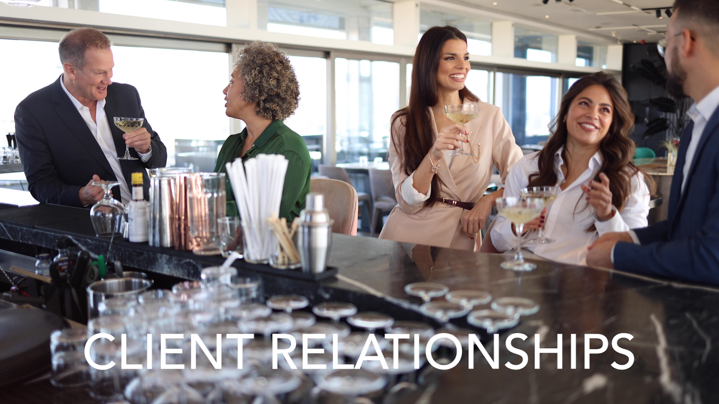 Group of people socializing at a bar, holding cocktails and engaging in conversation with a bartender behind the bar, in a modern restaurant or bar setting.