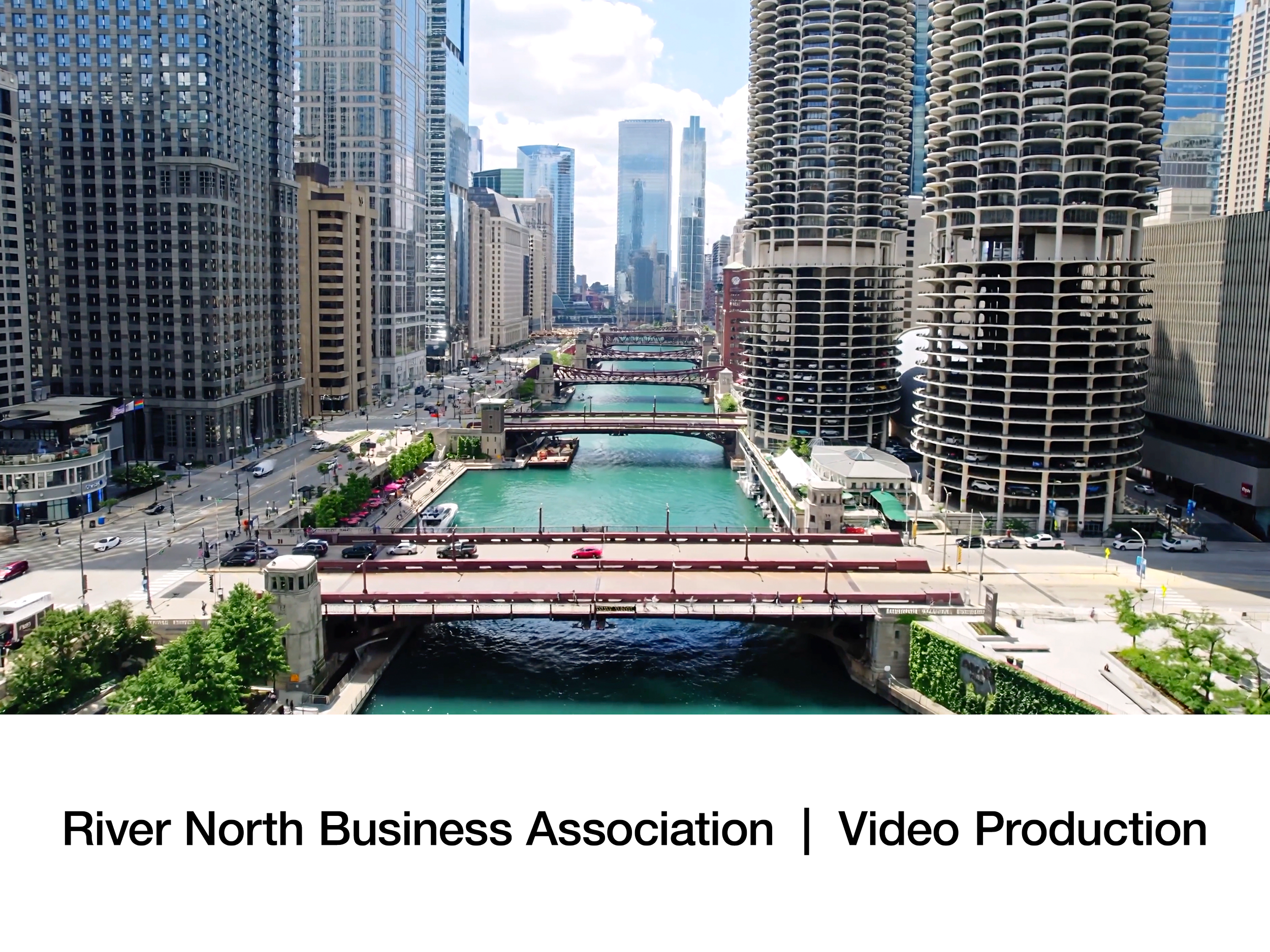 A wide shot of River North in Chicago with tall modern buildings, a river with greenish water, bridges crossing the river, and streets with cars and trees along the waterfront.