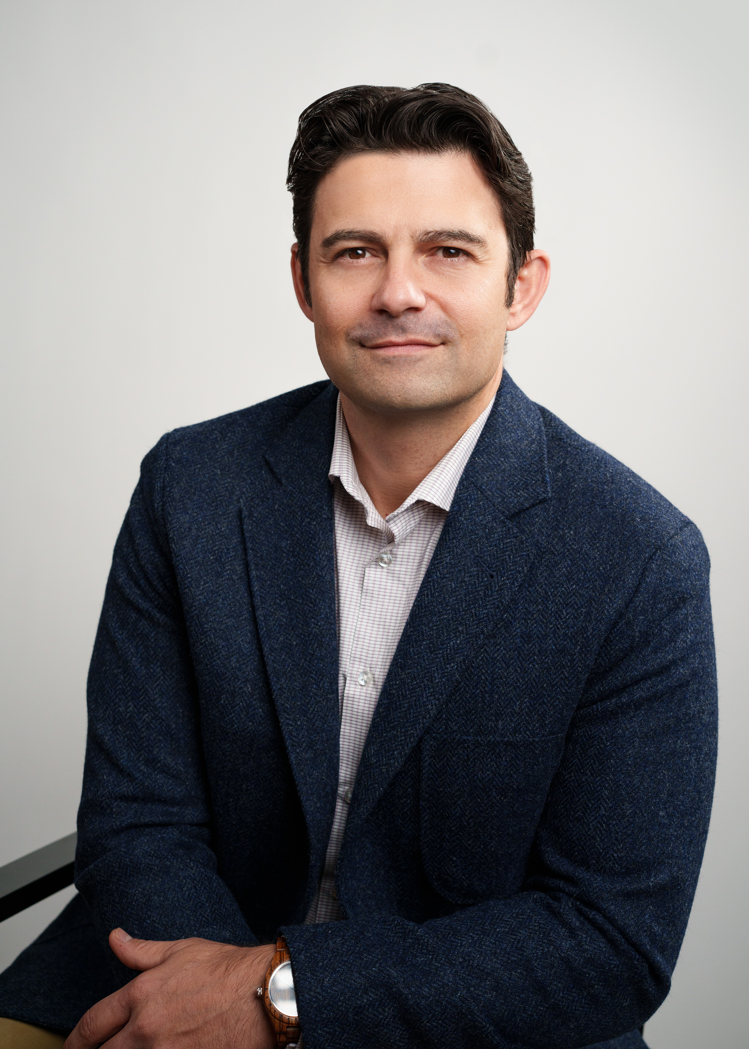 A man with dark hair, wearing a navy blazer over a white button-up shirt, sitting at a table with his left arm resting on it, against a plain white background.
