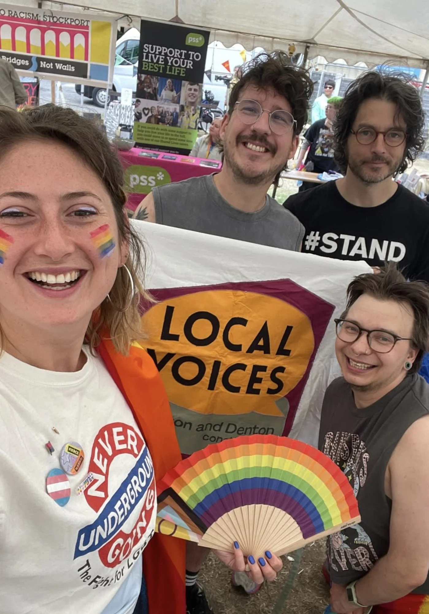 Four smiling people at a Pride stall holding a Local Voices banner and a rainbow fan