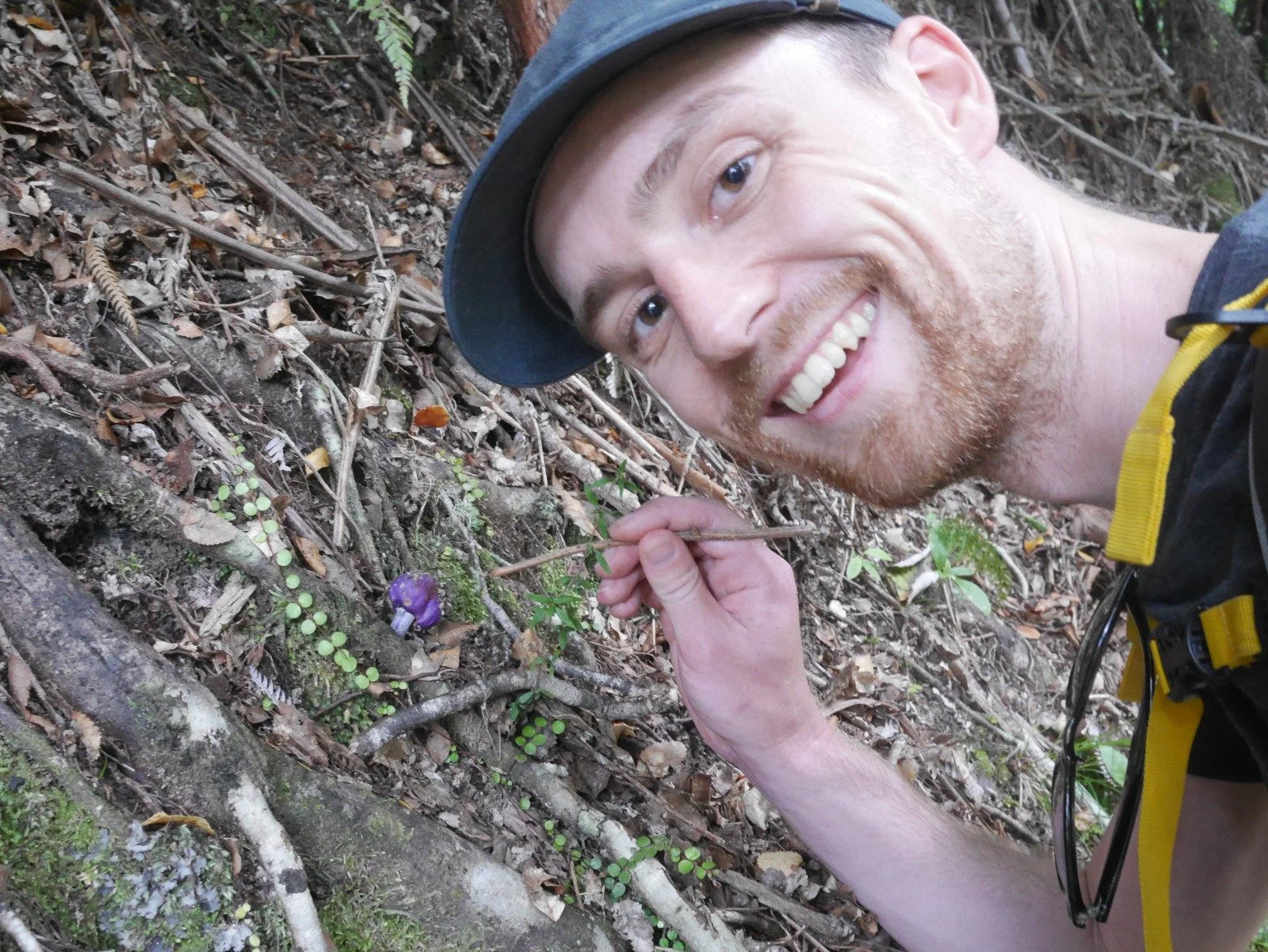 Tom smiling outdoors next to a small purple wildflower