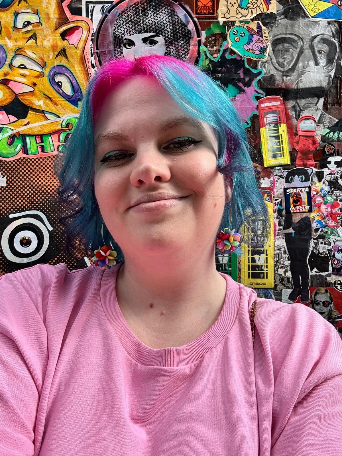 Greta, with pink and blue hair, smiling in front of a colourful mural
