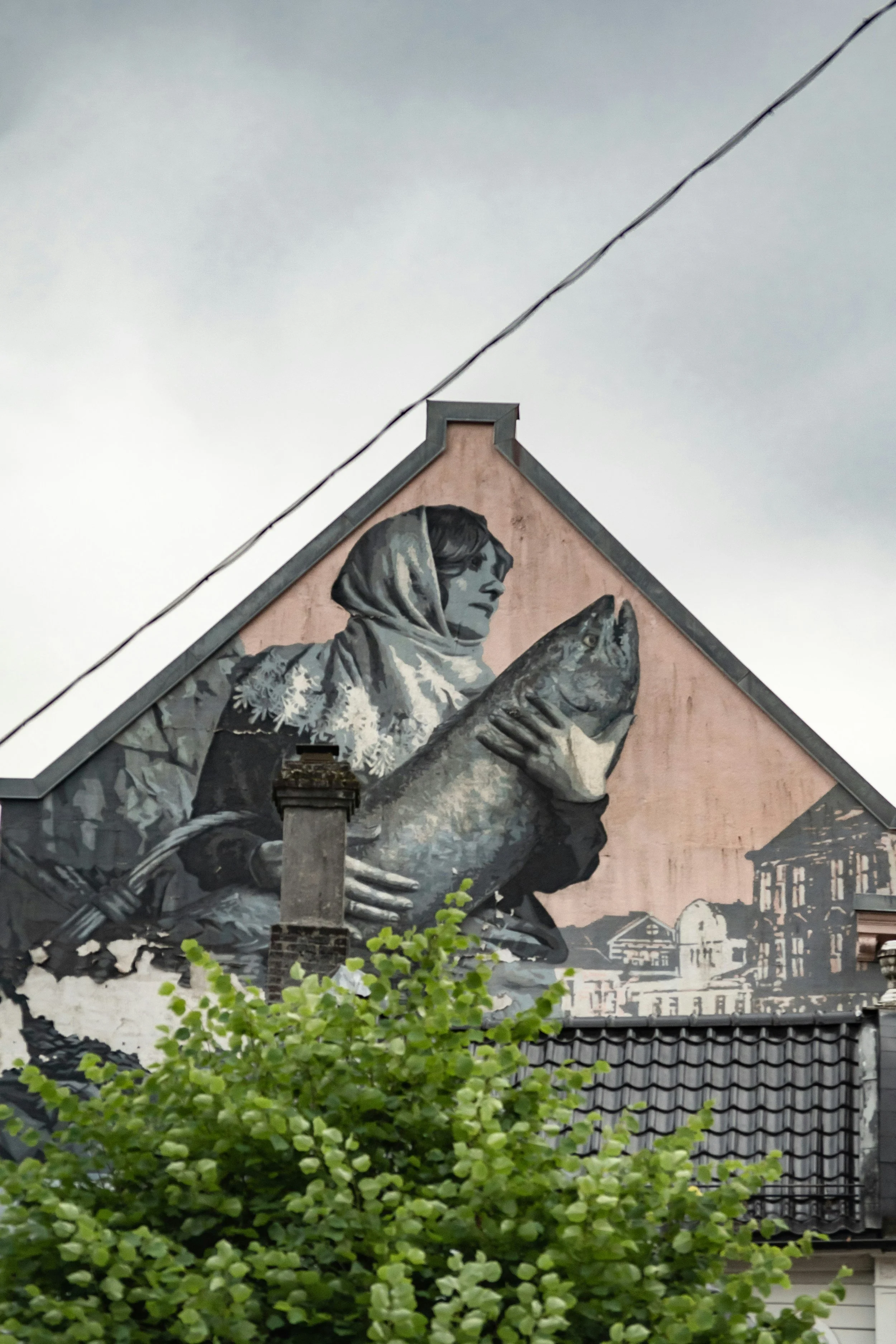 A mural painted on the side of a building shows a woman holding a fish, with a cityscape in the background. There is a tree with green leaves in the foreground.
