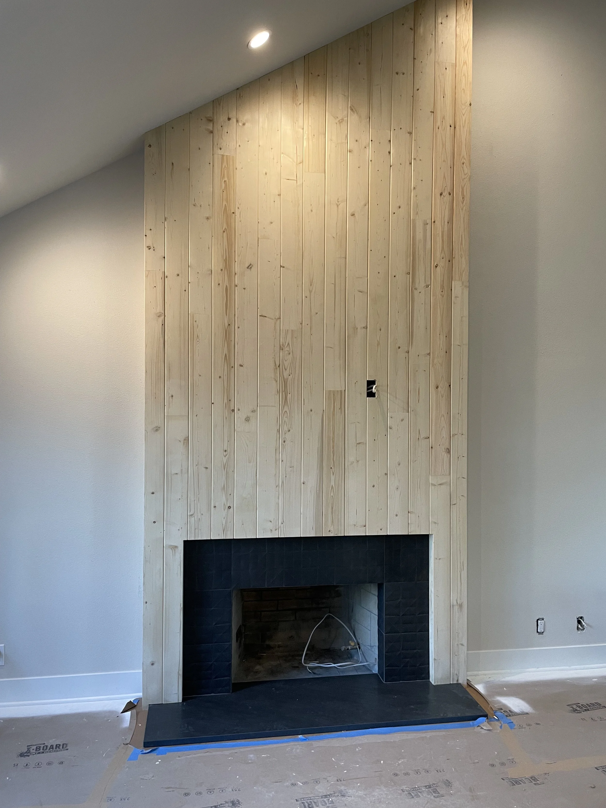 A fireplace with a black interior, surrounded by light-colored wooden paneling on an angled wall in a room under construction.