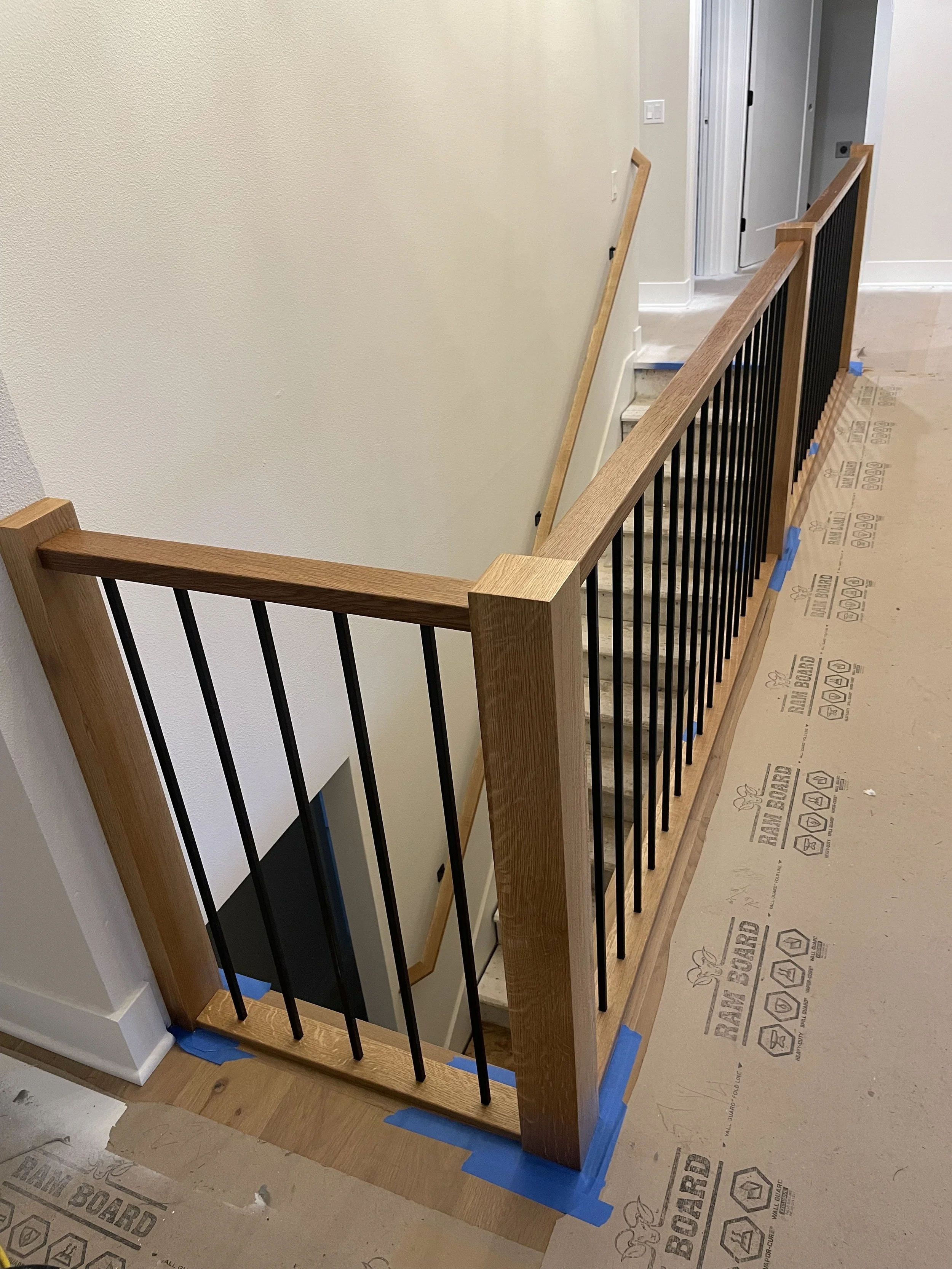View of a staircase construction with wooden handrail and black metal balusters, partially covered with painter's tape, in an interior under renovation.