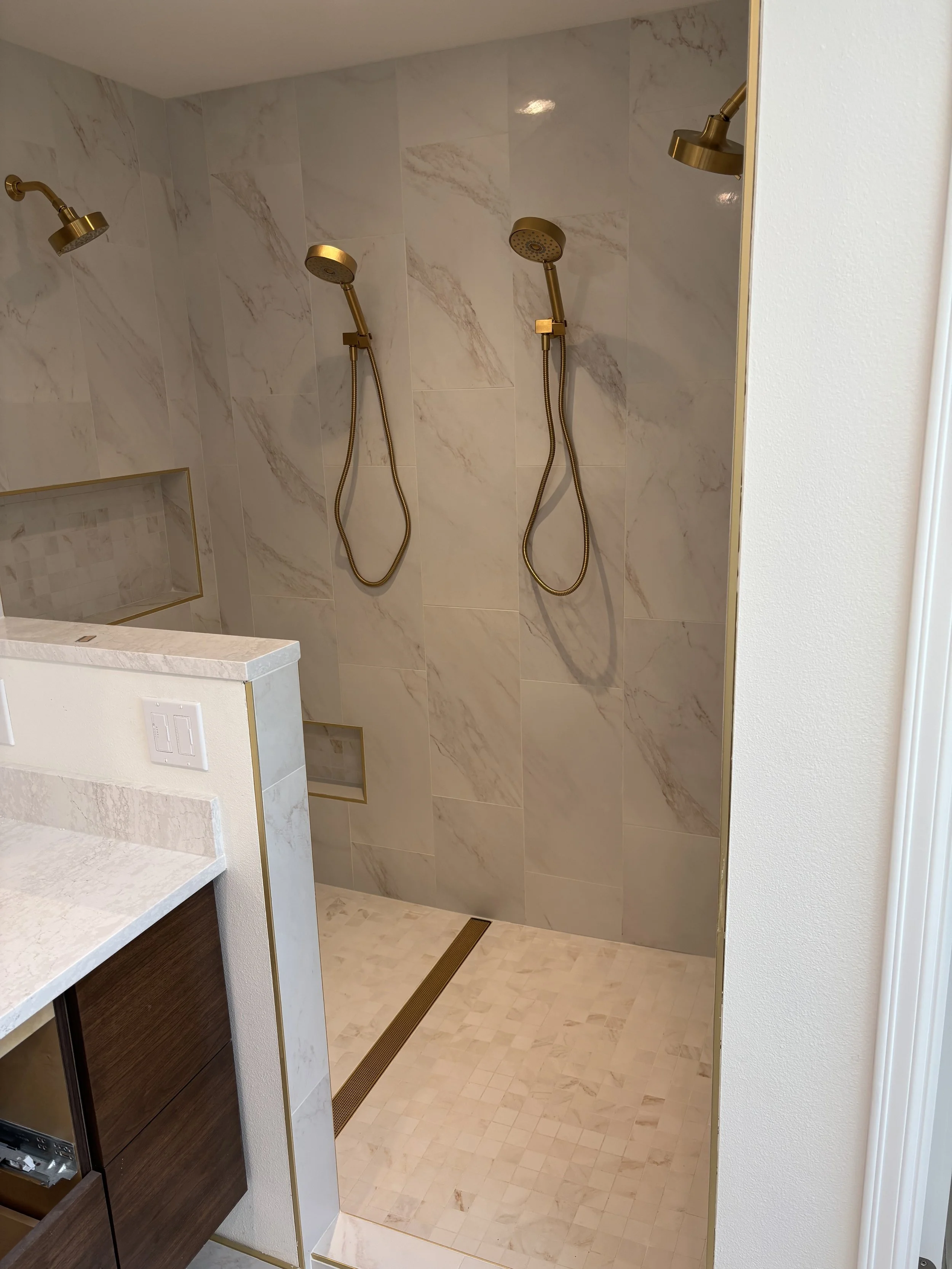 A walk-in shower area with three gold-colored showerheads on a marble wall and a drainage grate on the beige tiled floor.