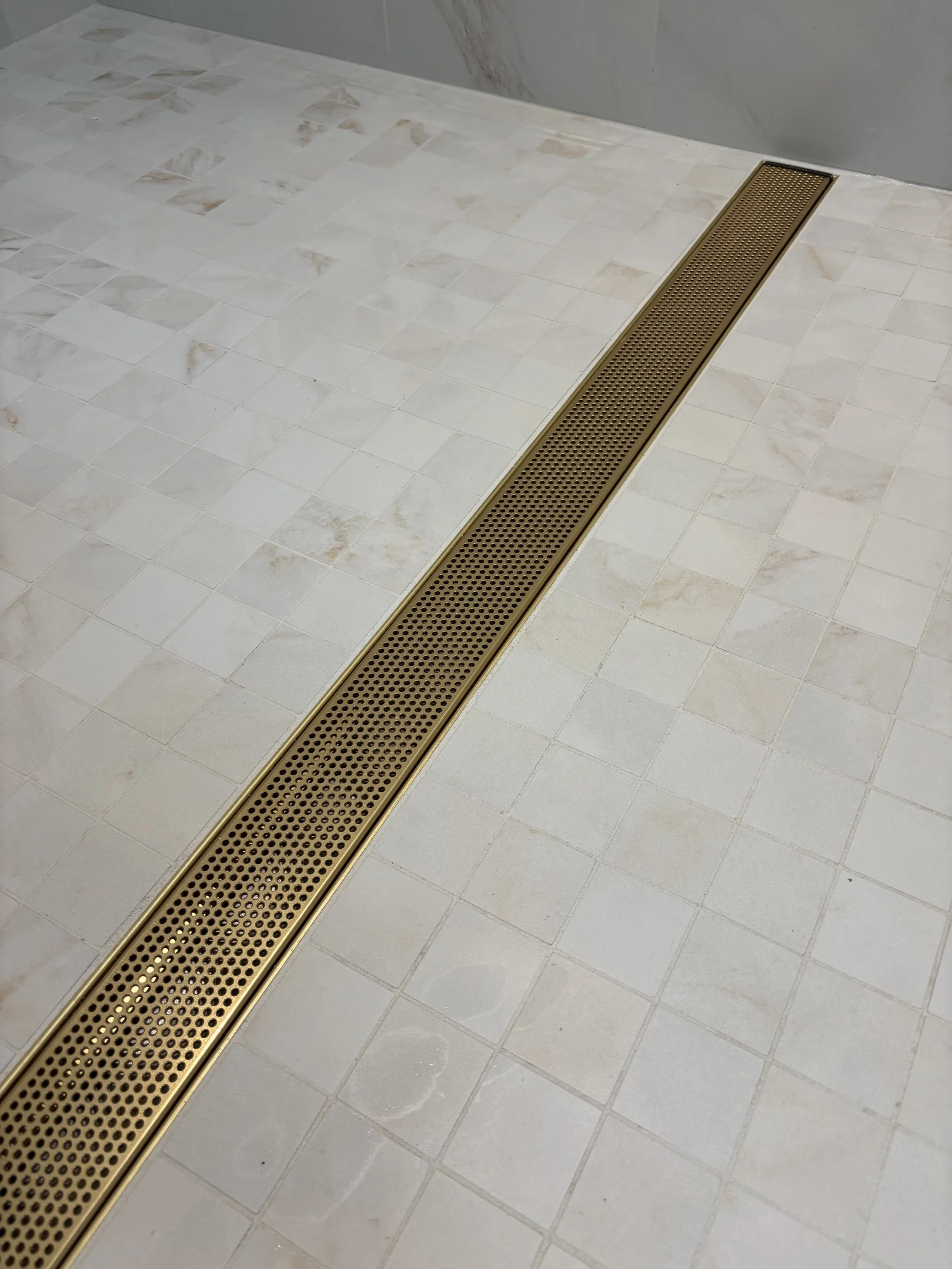 A beige-colored floor drain cover installed in a white tile floor, with the surrounding tiles having a marble pattern, and a gray wall in the background.