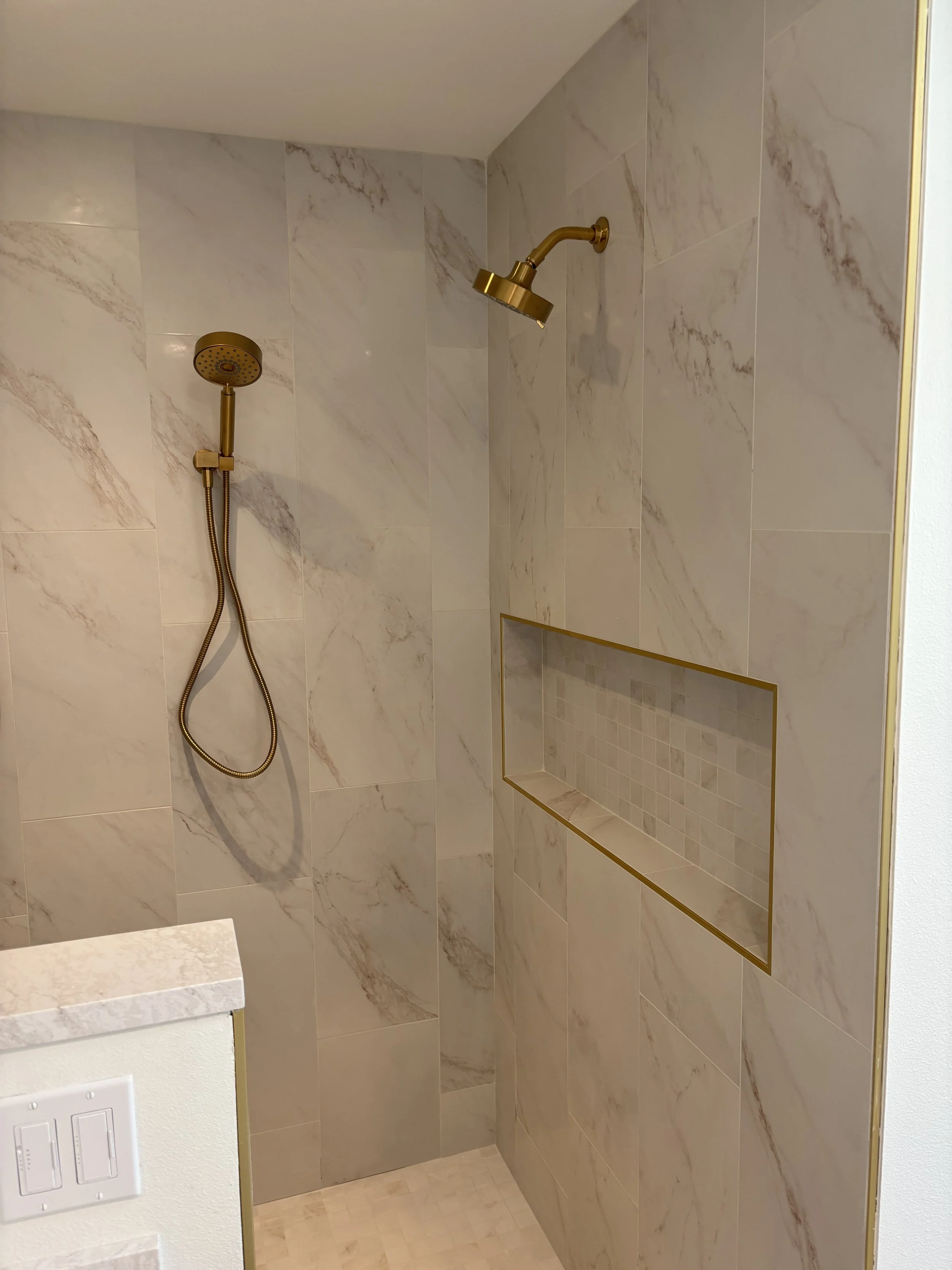 Shower with marble tiles, gold fixtures including a showerhead and handheld spray, built-in niche with small tiles, and a white wall with a light switch.