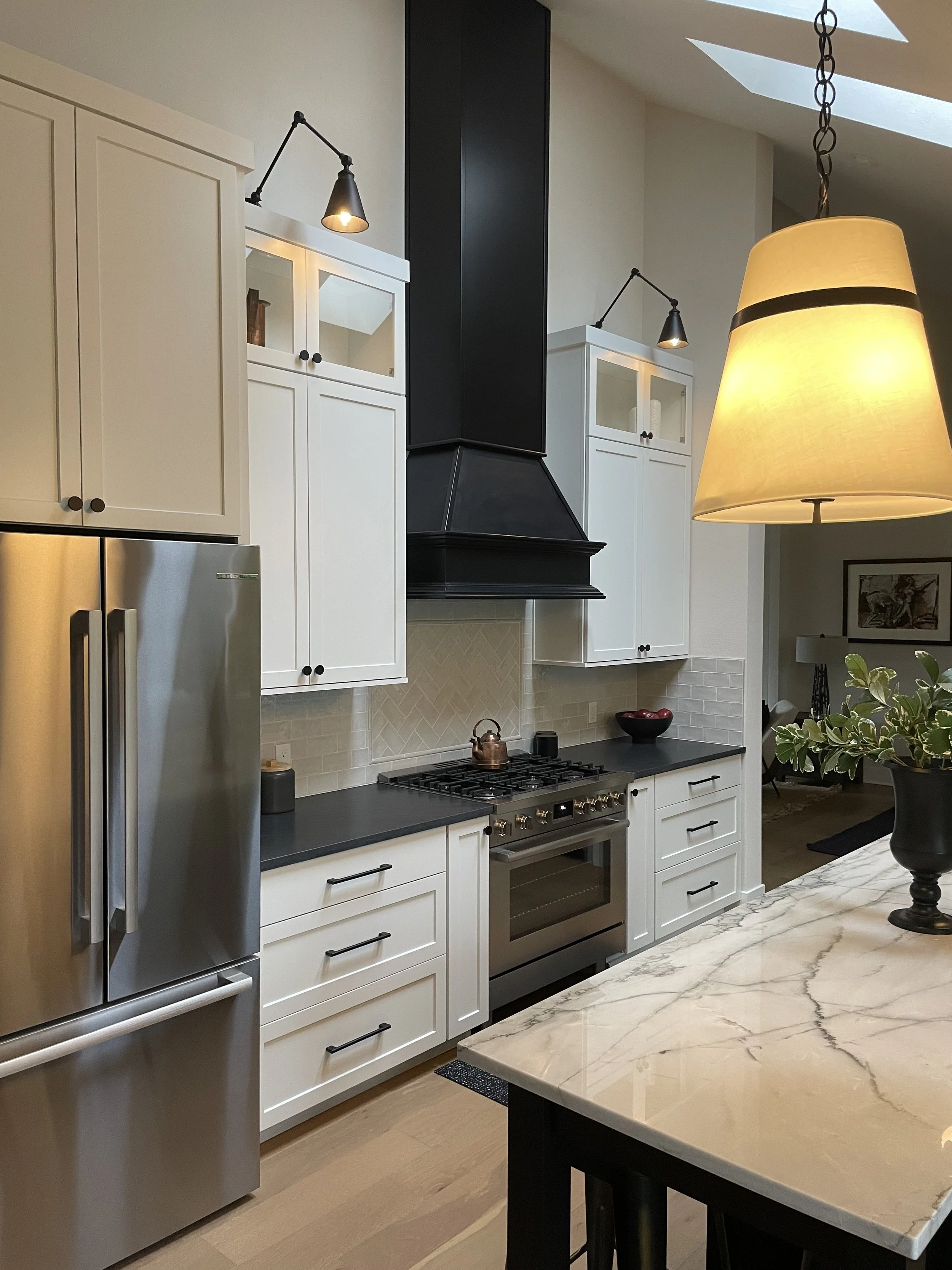 Modern kitchen with white cabinets, stainless steel refrigerator, black stovetop, black range hood, marble countertop, and a hanging lamp. A bowl and plants are on the counter, with ceiling skylights in the background.