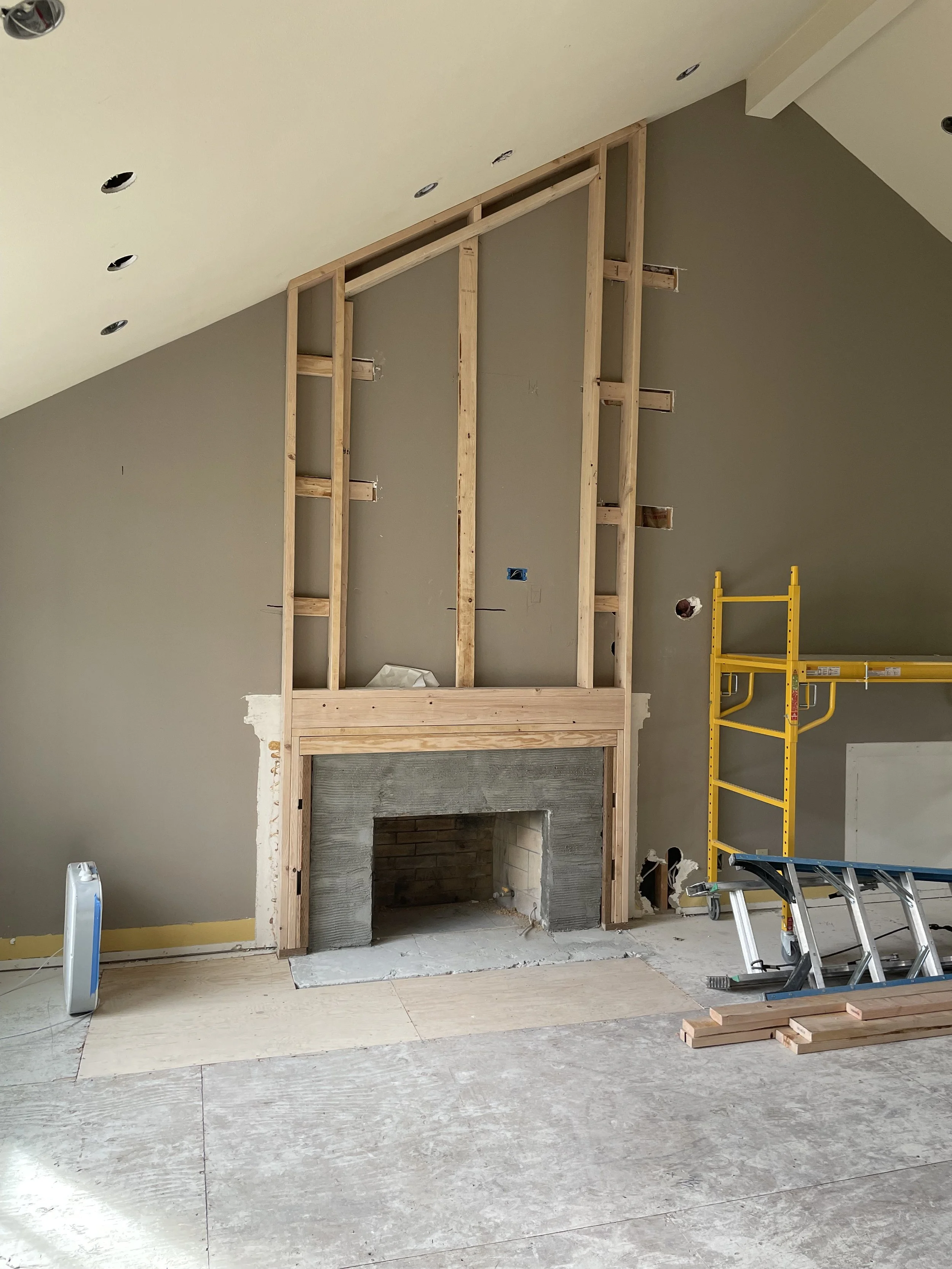 Interior of a room under construction with a partially built wooden frame around a fireplace, a stepladder, a yellow scaffold, and construction materials on the floor.