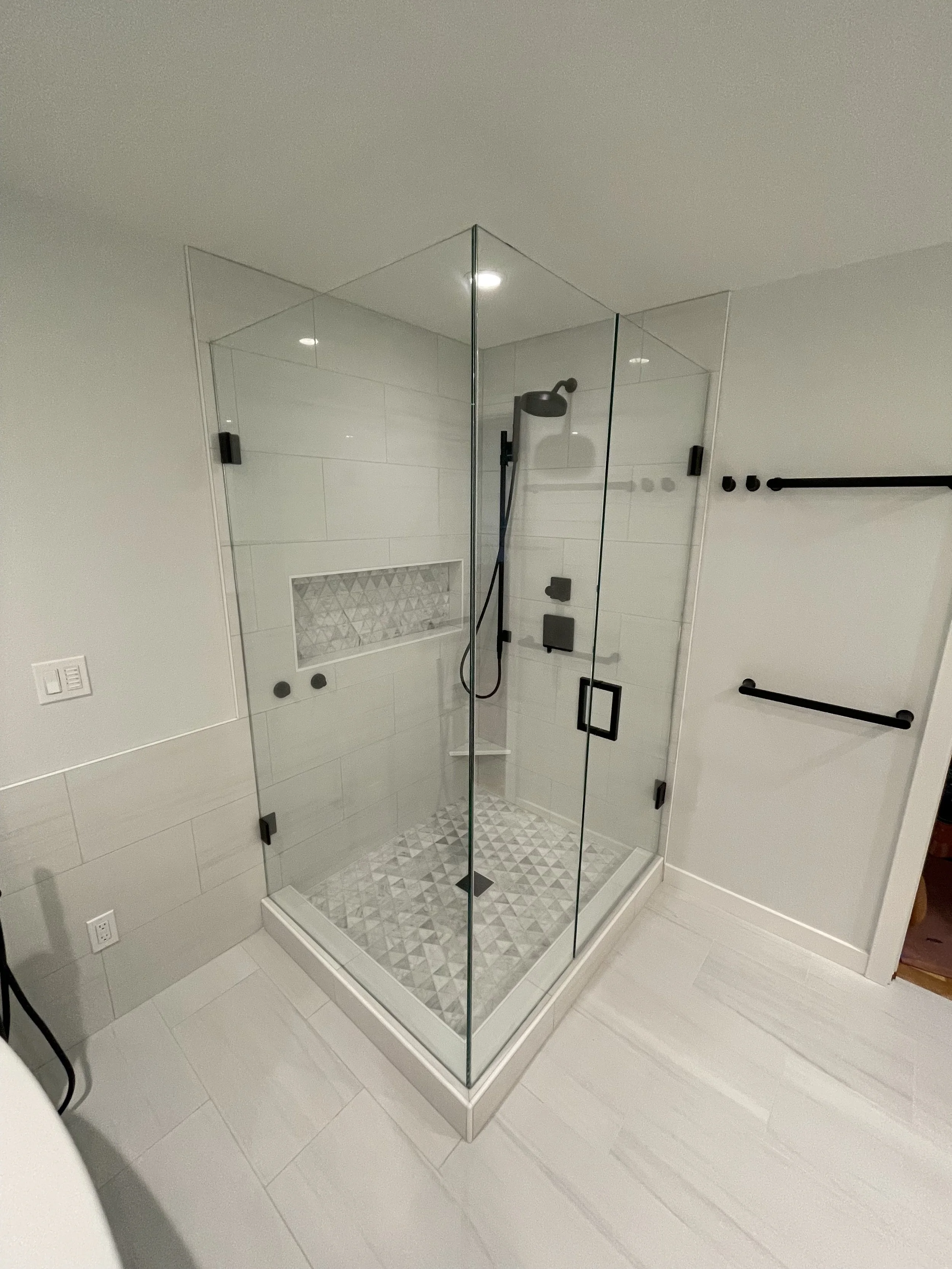 A modern glass shower enclosure with black fixtures in a white-tiled bathroom.