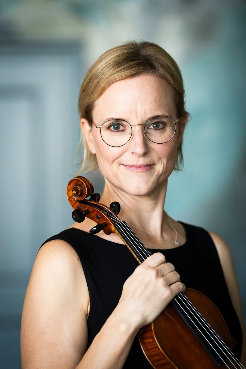 A woman with short blonde hair, glasses, and a black sleeveless top holding a violin.